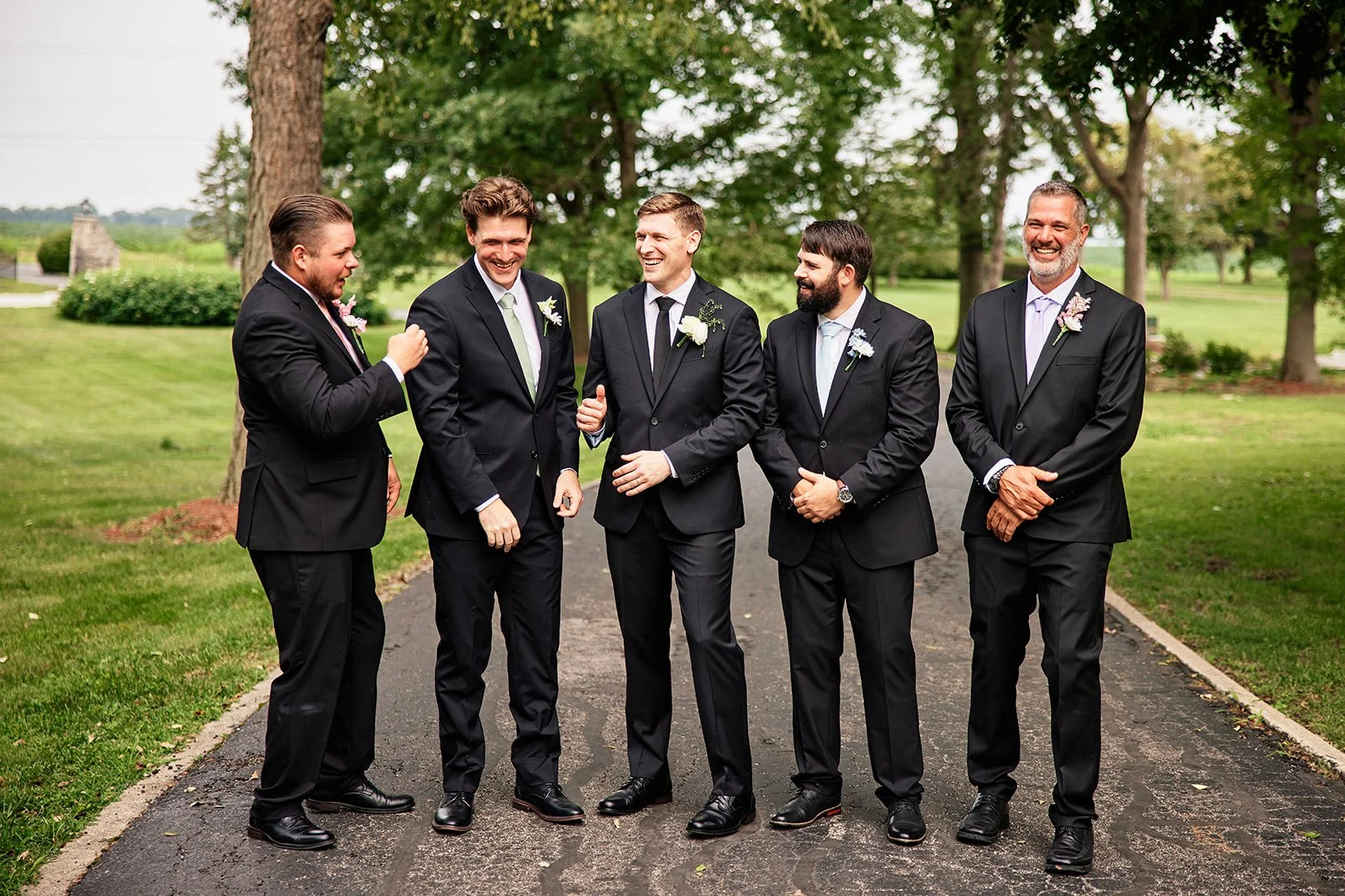 Five men in black suits standing outdoors on a paved path, engaged in conversation and laughing, with trees and grass in the background. Groomsmen, best man and groom. 
