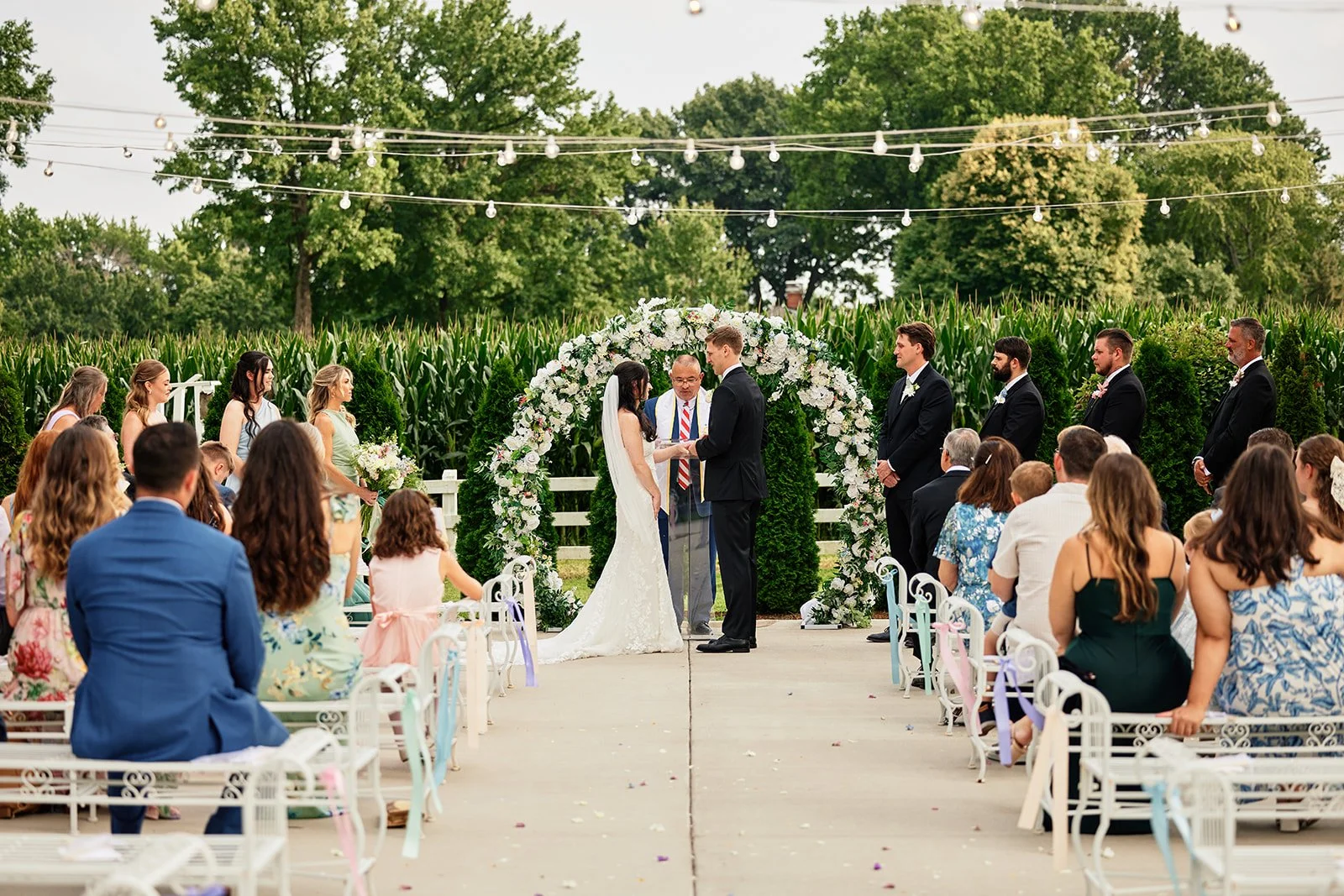 A wedding ceremony outdoors with a bride and groom exchanging vows under a floral arch. Guests seated on white benches observe, with green trees and cornfields in the background, and string lights hanging overhead.