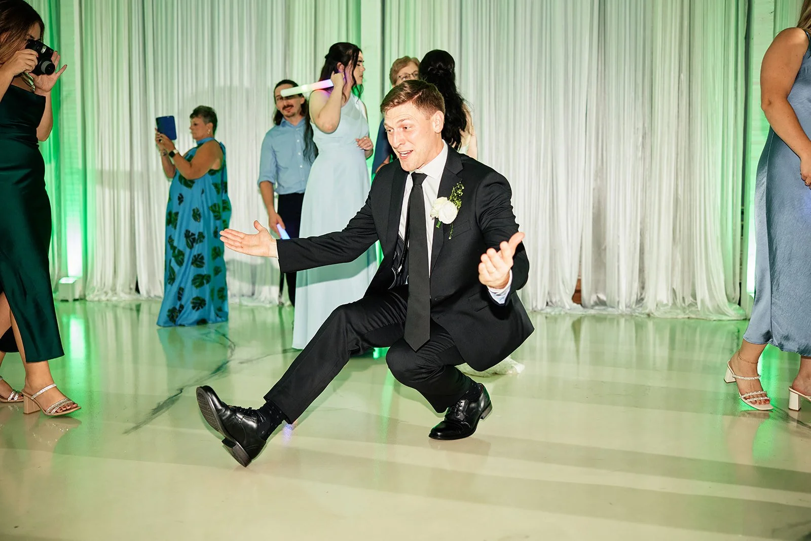 A man (groom) in a black suit and tie kneeling on the dance floor with arms outstretched, smiling, at a wedding reception. People in formal attire are dancing and taking photos in the background, with a backdrop of white curtains and green lighting.