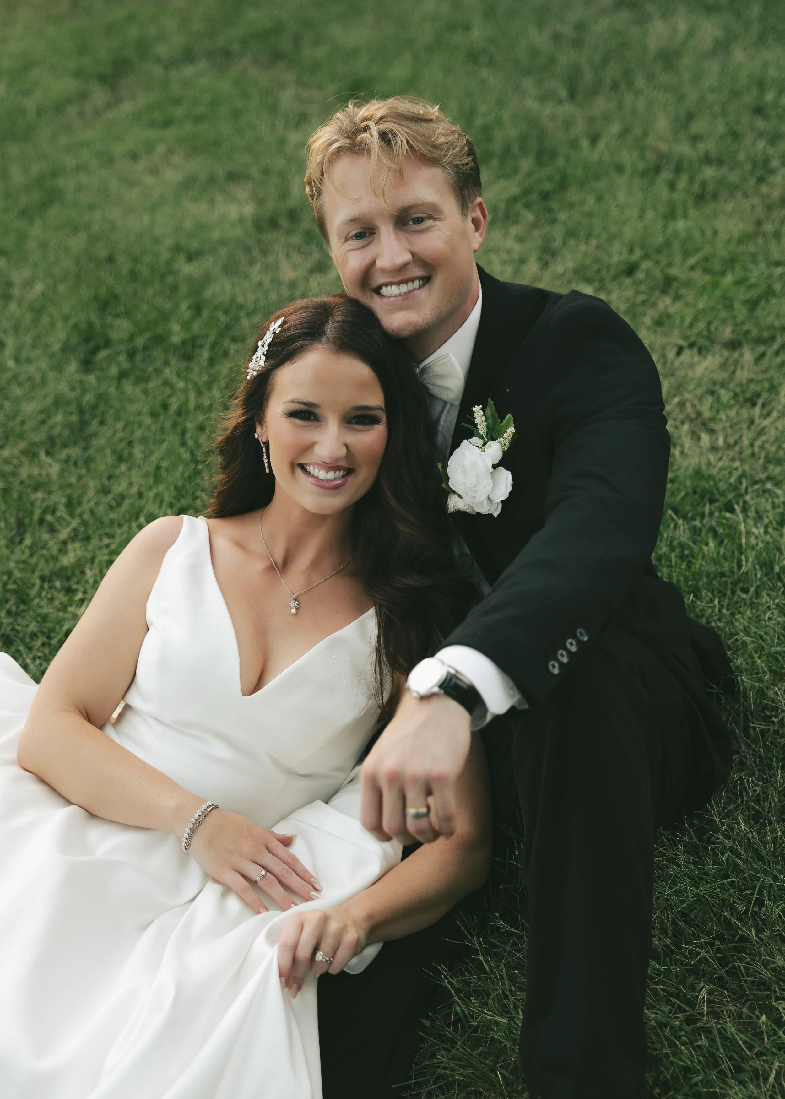 A smiling bride and groom lying on grass, the bride in a white wedding dress with jewelry, and the groom in a black tuxedo with a white boutonniere, holding hands.