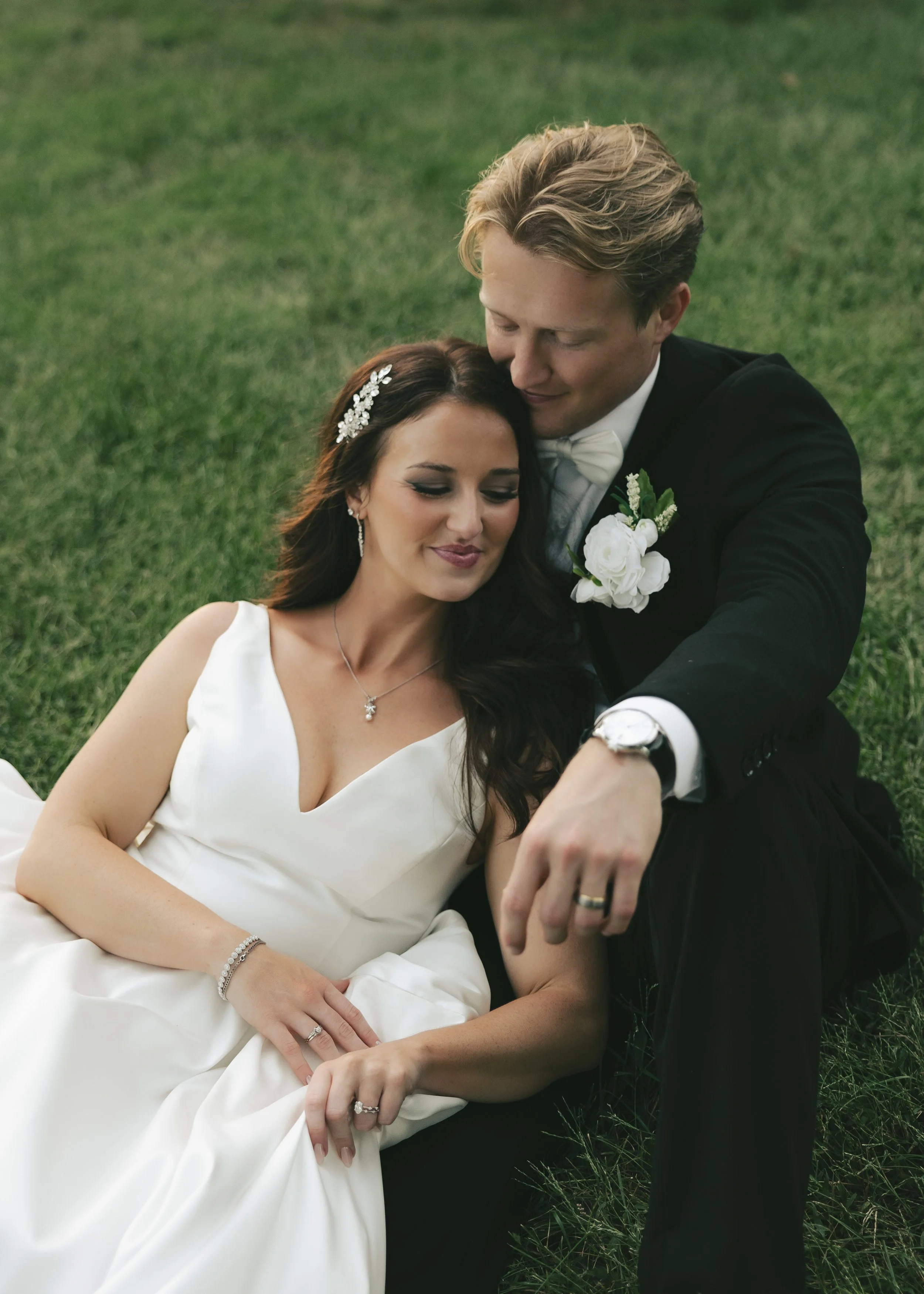 A bride and groom sitting on the grass, with the groom gently holding the bride's hand.