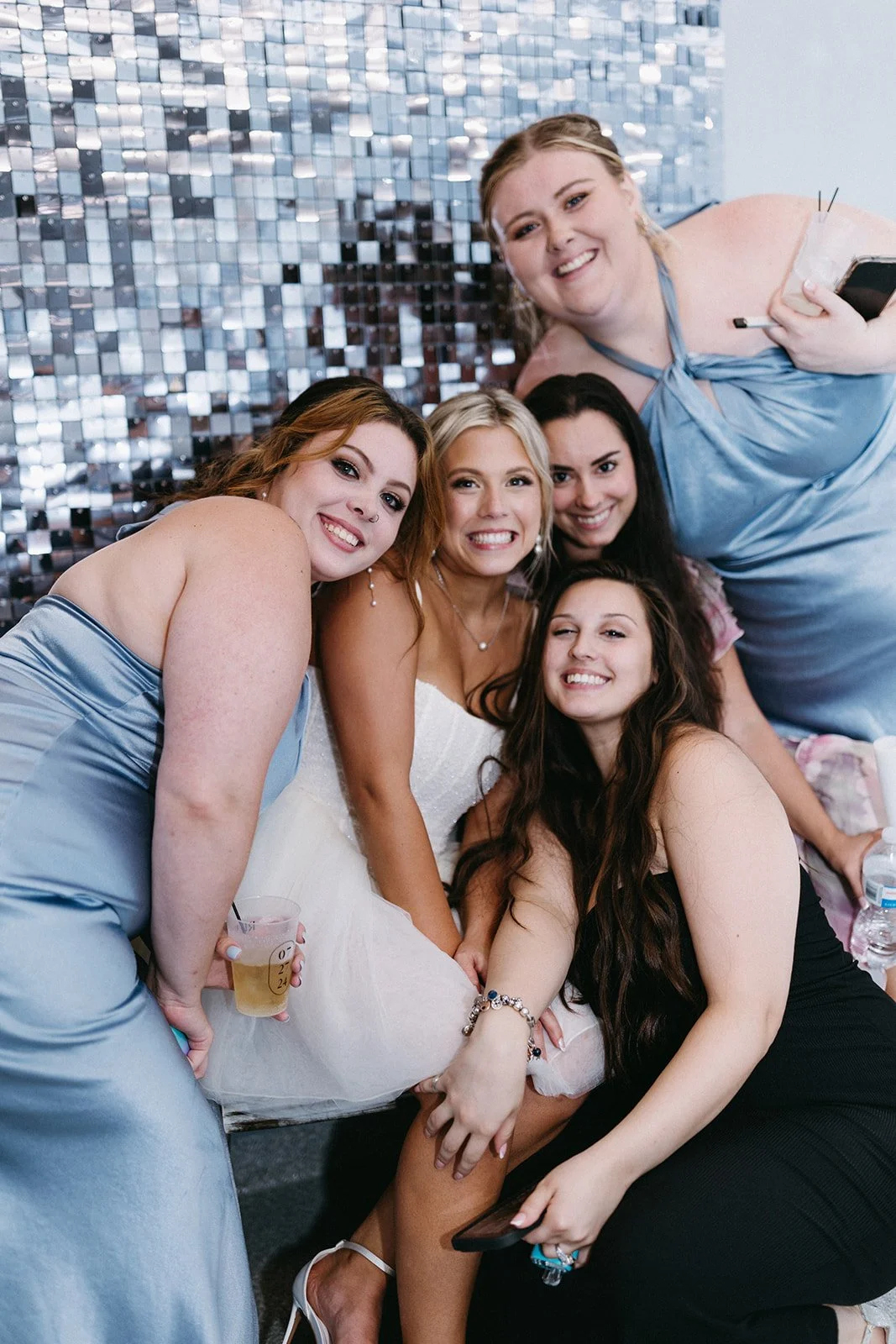 Group of five women smiling, dressed in elegant attire, at a celebration or event, with a shiny, sequined backdrop.