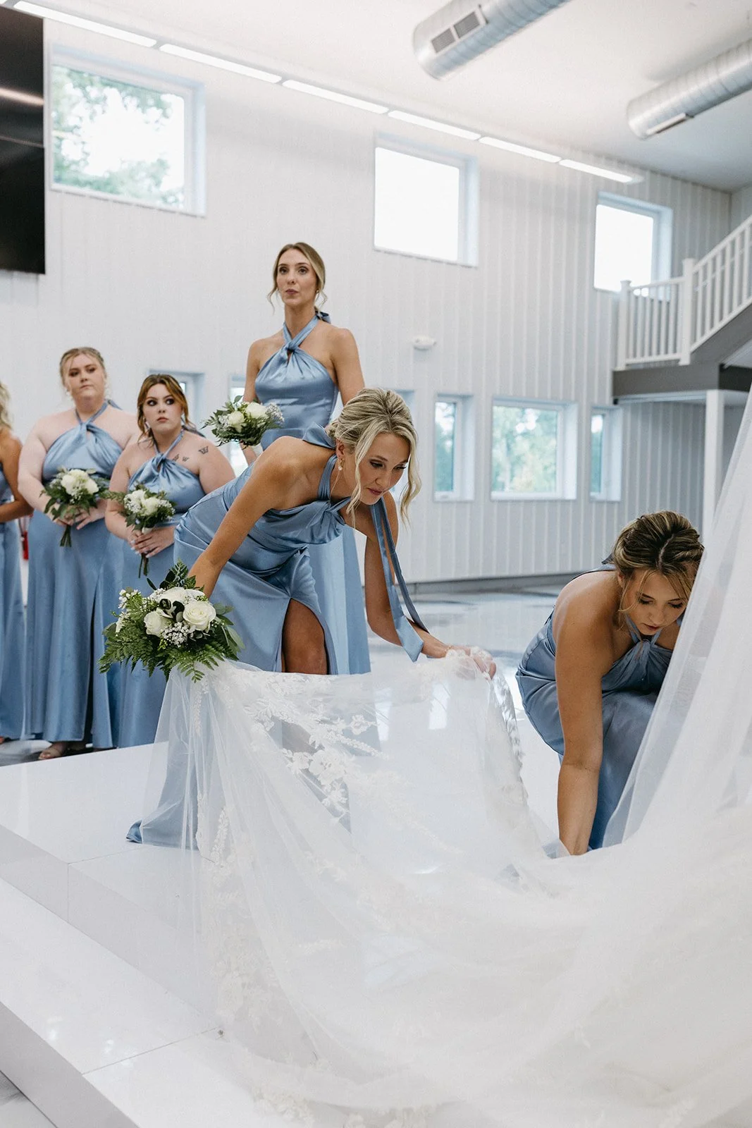 Bridesmaids in blue dresses participating in a wedding ceremony, with some placing a wedding dress into a white casket inside a bright hall with large windows.