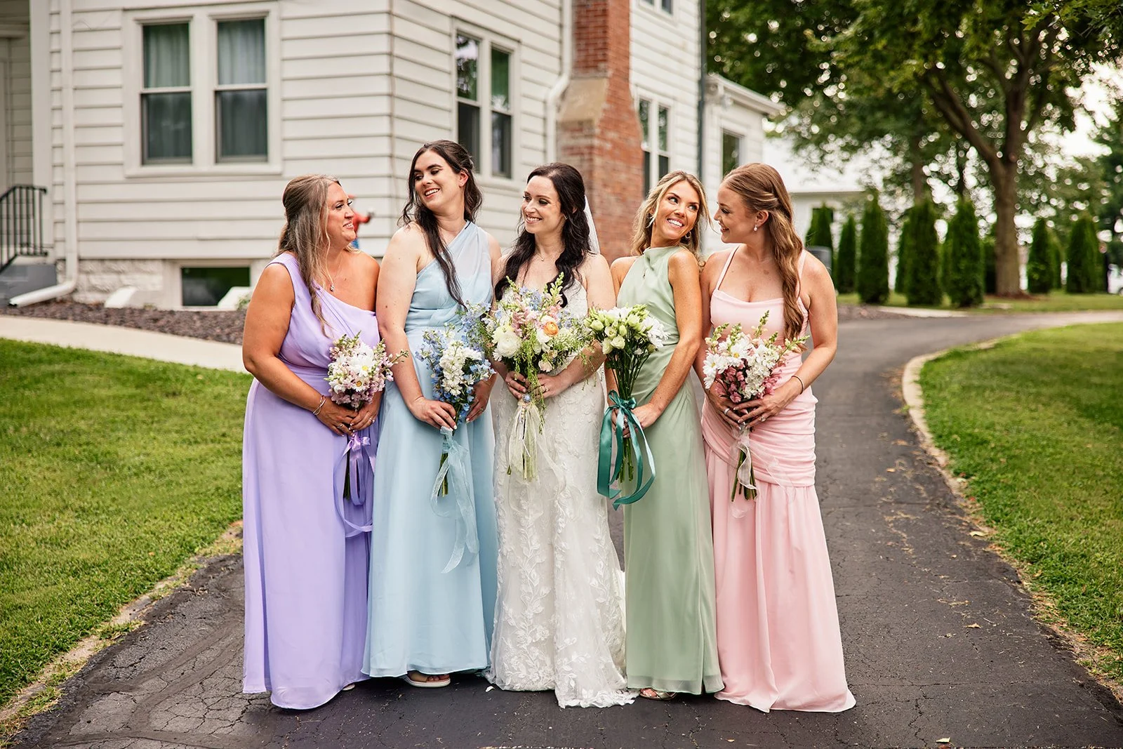 Group of five women in colorful dresses, standing outdoors on a curved asphalt pathway, holding bouquets, with a residential house and trees in the background, celebrating a wedding. Bridesmaids in pastel dresses.