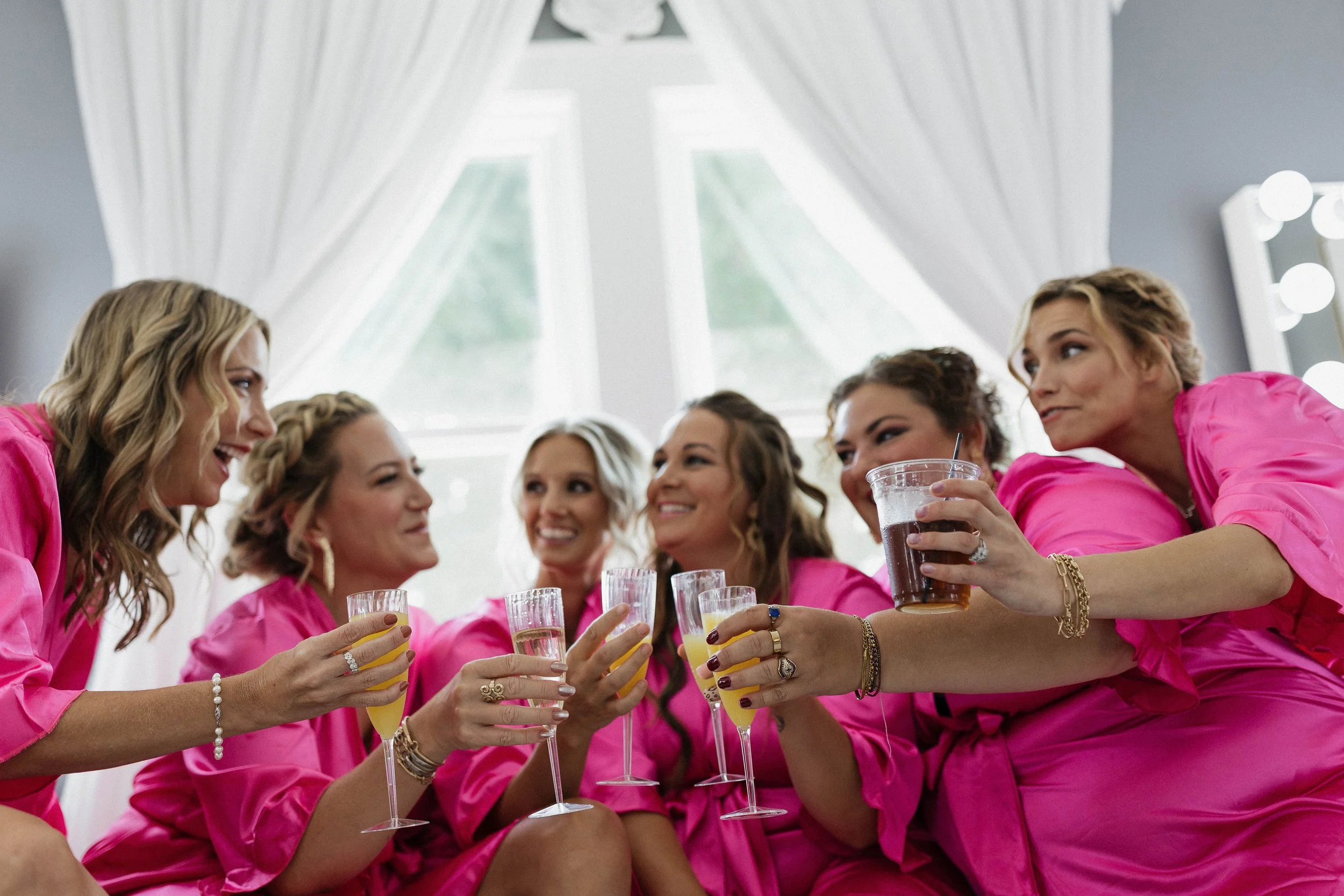 A group of six women in pink robes sitting together, smiling and toasting with drinks in a bright room with large windows and white curtains.