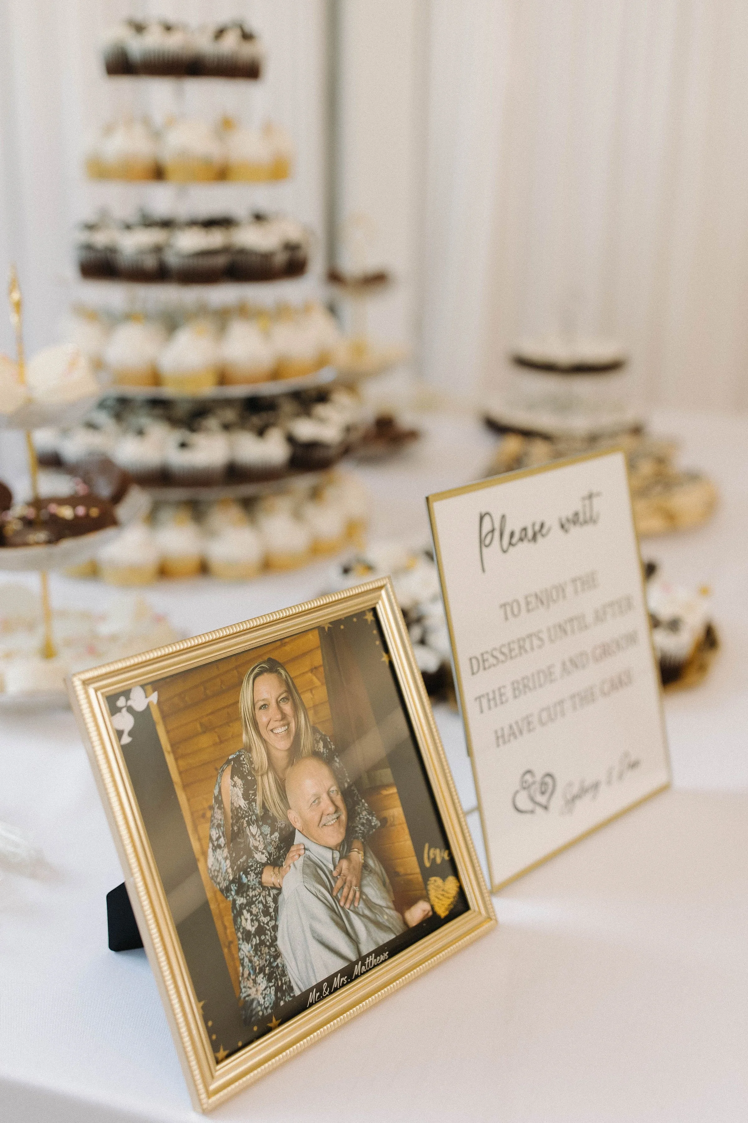 A framed photo of a smiling woman and an elderly man sitting together, with a dessert table featuring cupcakes and pastries in the background, and a sign with wedding instructions.