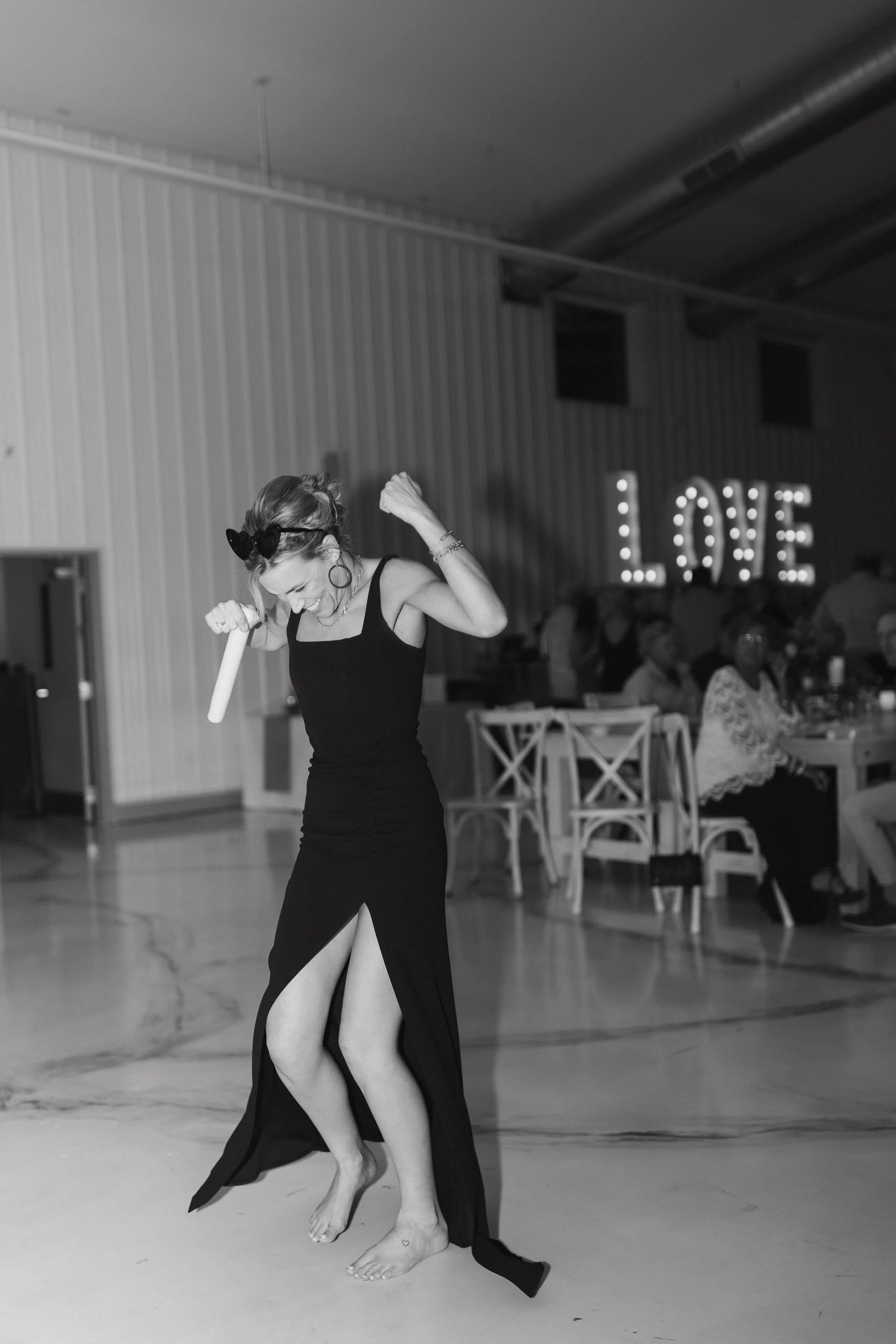 A woman in a black dress dancing barefoot at a celebration, with a large illuminated 'LOVE' sign in the background.