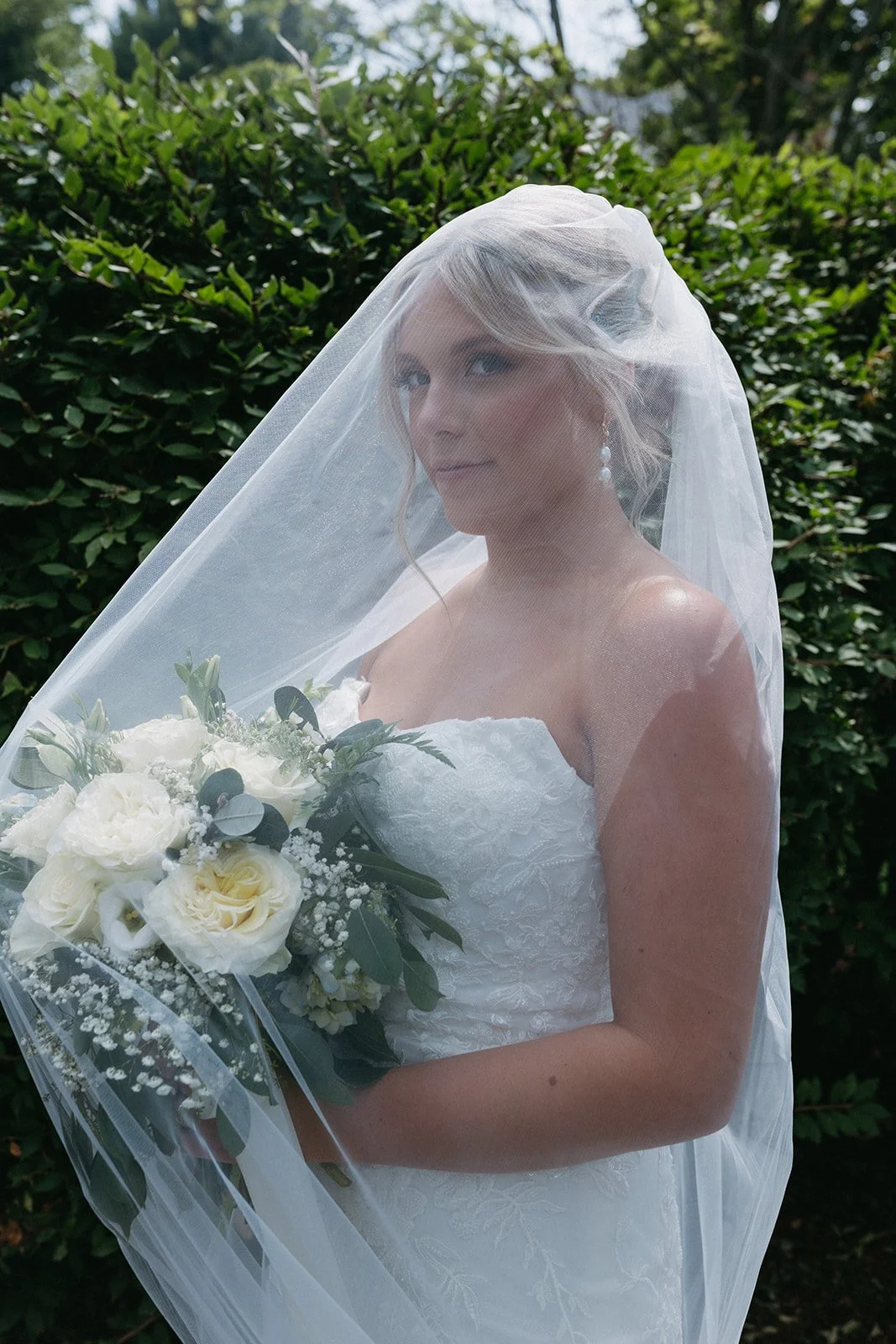Bride in a white wedding dress holding a bouquet of white flowers, with a veil covering her head, outdoors against green foliage.