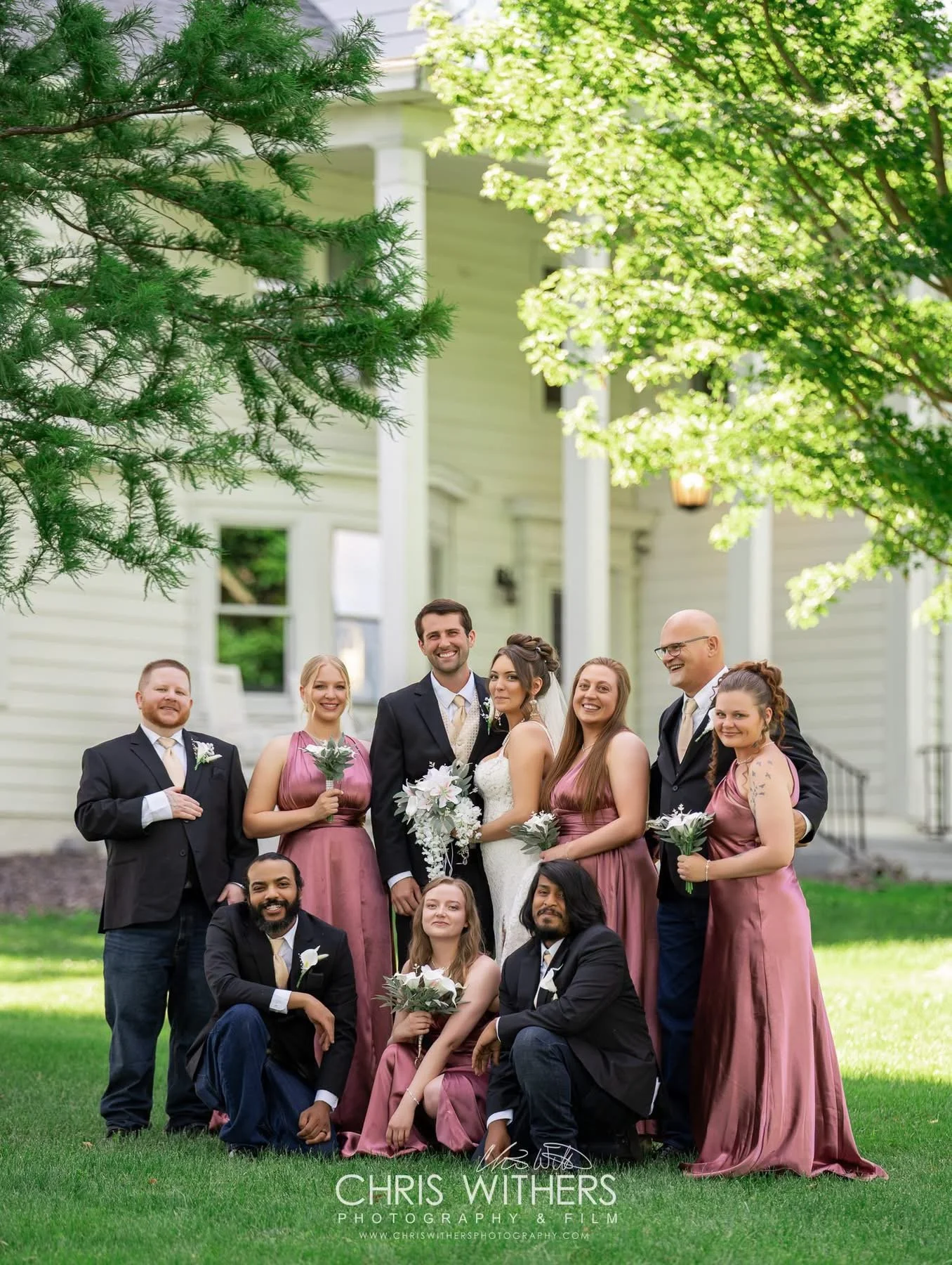 A group of wedding people standing on a lawn in front of a white house, with some trees and greenery around.