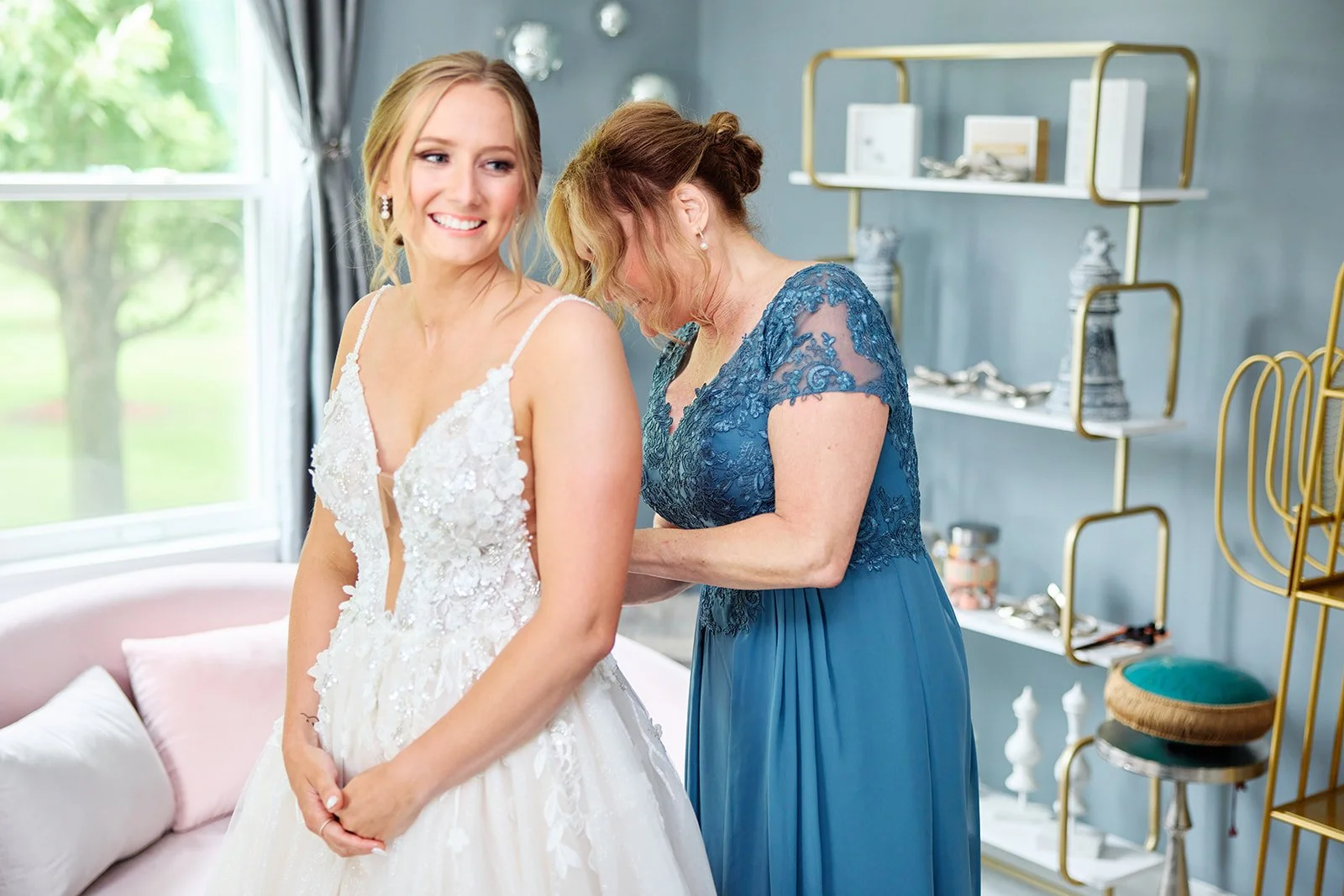 A bride in a white lace wedding dress smiling as her mother adjusts her gown in a bright, modern room with decorative shelving and a window showing greenery outside.