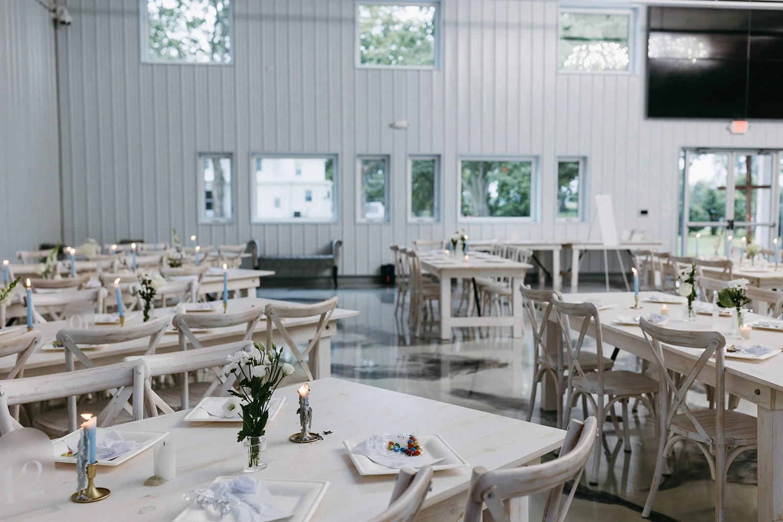 Decorated banquet hall with white tables and chairs, floral centerpieces, candles, and a modern rustic indoor setting.