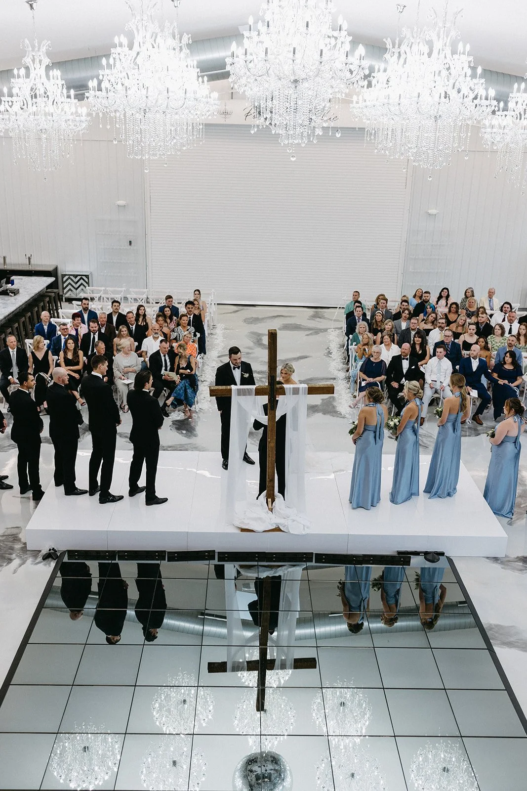 Wedding ceremony with a cross and officiant on a white stage, surrounded by bridesmaids in blue dresses and groomsmen in black suits, guests seated in rows in a spacious, elegant hall with crystal chandeliers.