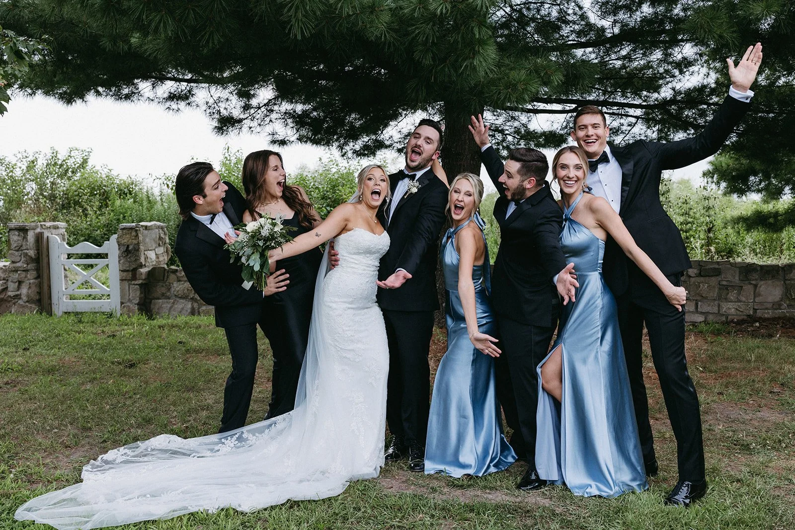 A wedding party celebrating outdoors under a large tree, with the bride in a white wedding gown and the groom in a black tuxedo, surrounded by friends and bridesmaids in matching blue dresses and groomsmen in black suits, all smiling and laughing.