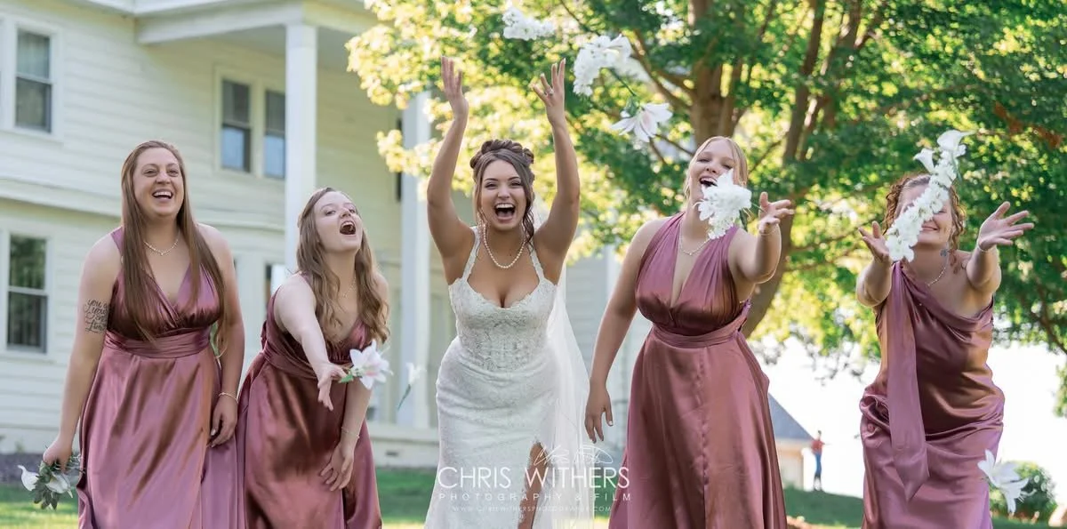 Bride in white wedding dress celebrating with her bridesmaids, all in matching mauve dresses, outdoors under a large tree on a sunny day.