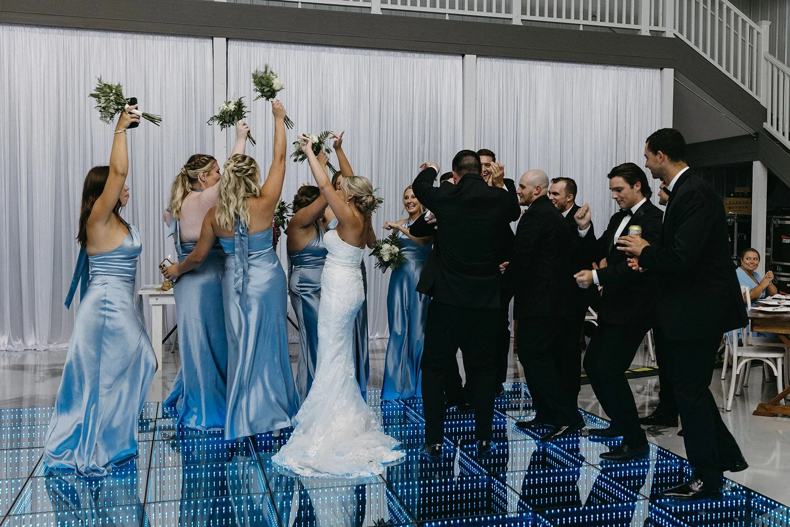 A wedding reception with women in blue bridesmaid dresses and a bride in a white gown dancing and celebrating, while men in black tuxedos dance and have fun.