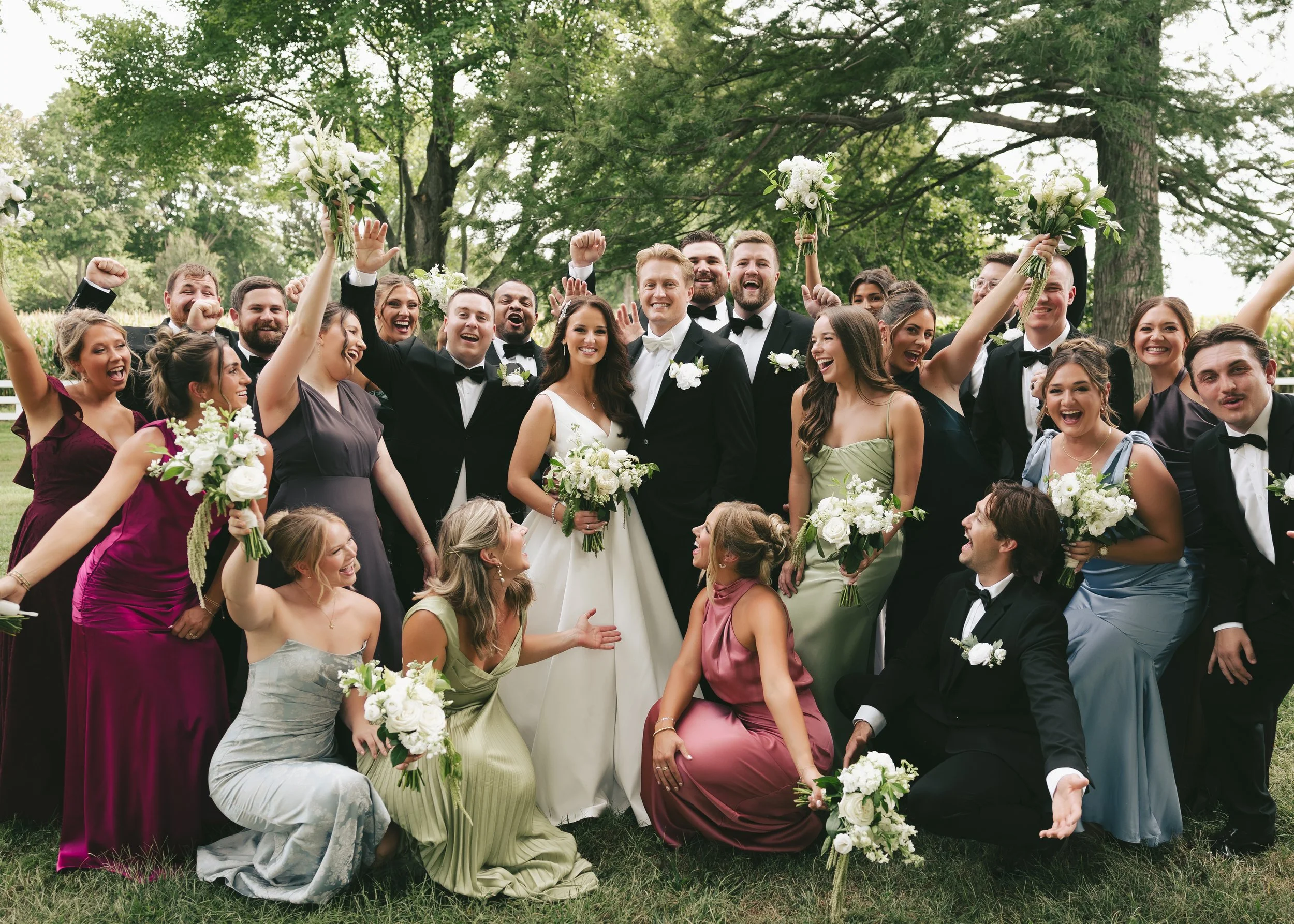 A large group of wedding guests, dressed in formal attire, celebrating outdoors at the wedding venue Pole Barn Chic with trees in the background. They are smiling, cheering, and holding bouquets of flowers. Central IL