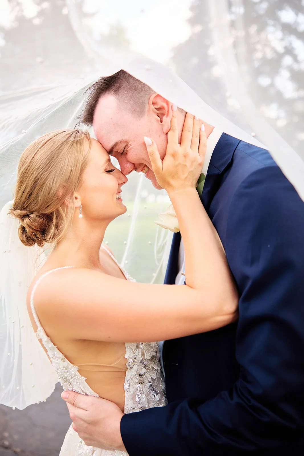 A bride and groom smiling closely under a wedding veil, holding each other's faces, with their foreheads touching during their wedding.