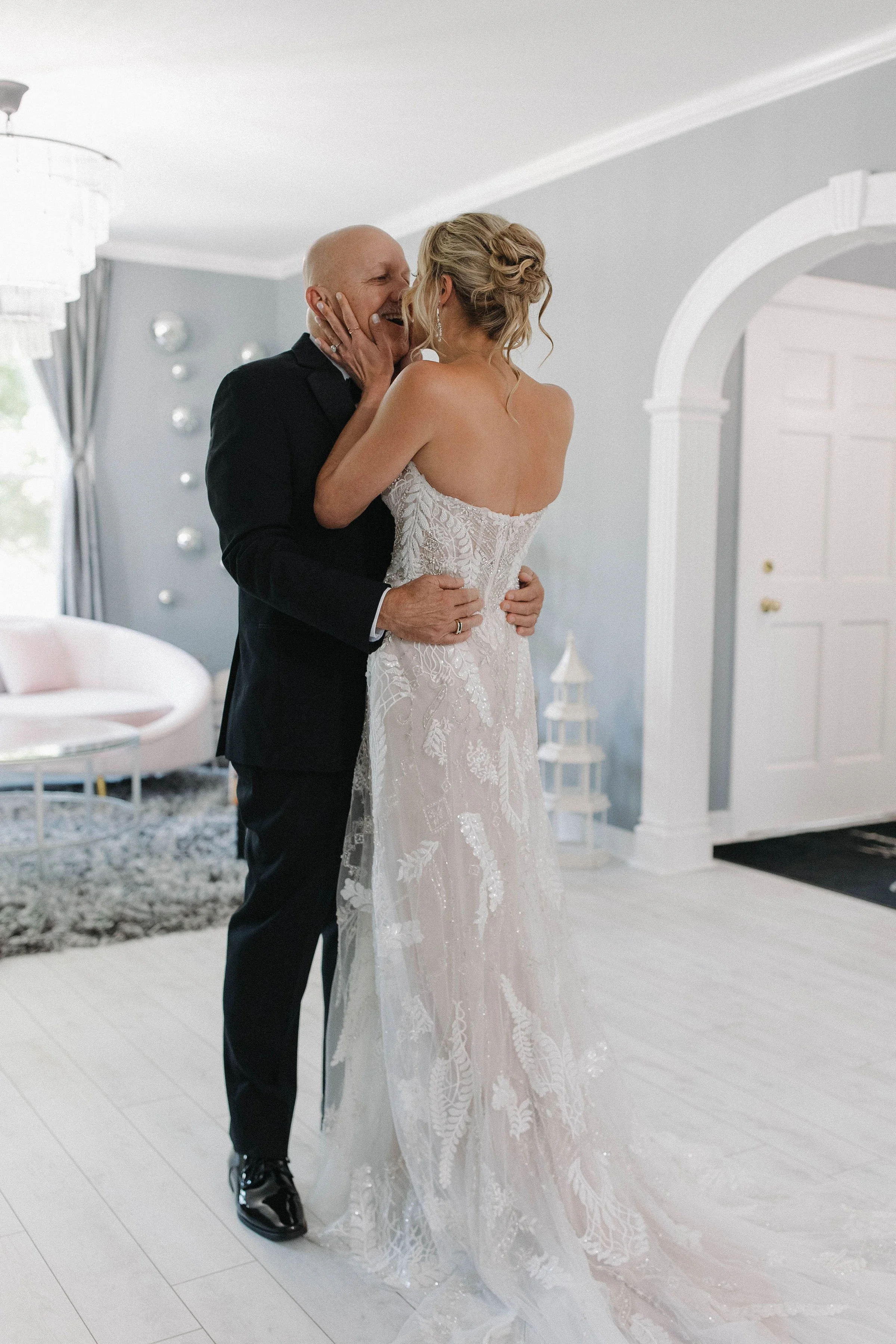 A bride and an older man, possibly her father, share an emotional moment inside a bright, modern home. The bride is in a strapless wedding gown, and the man is in a black suit. They are touching faces and smiling, surrounded by minimalist decor inclu