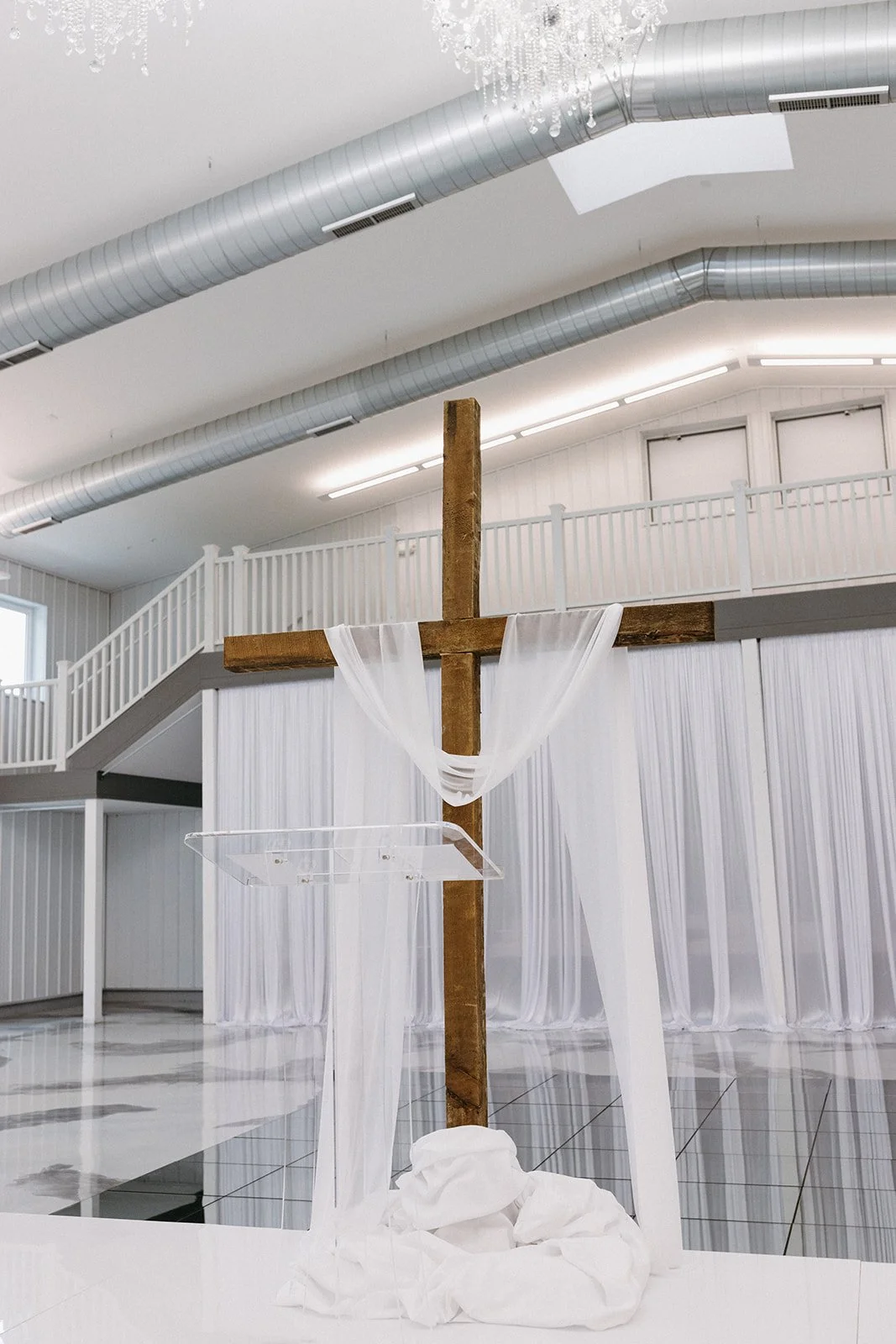Wooden cross draped with white fabric, set on a white cloth at the front of a church or chapel with white walls and a balcony railing.