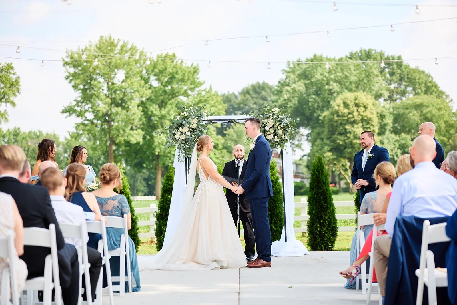 Bride and groom holding hands during outdoor wedding ceremony with guests seated on white chairs, officiant standing behind them, decorated arch with flowers, large green trees and string lights in the background.