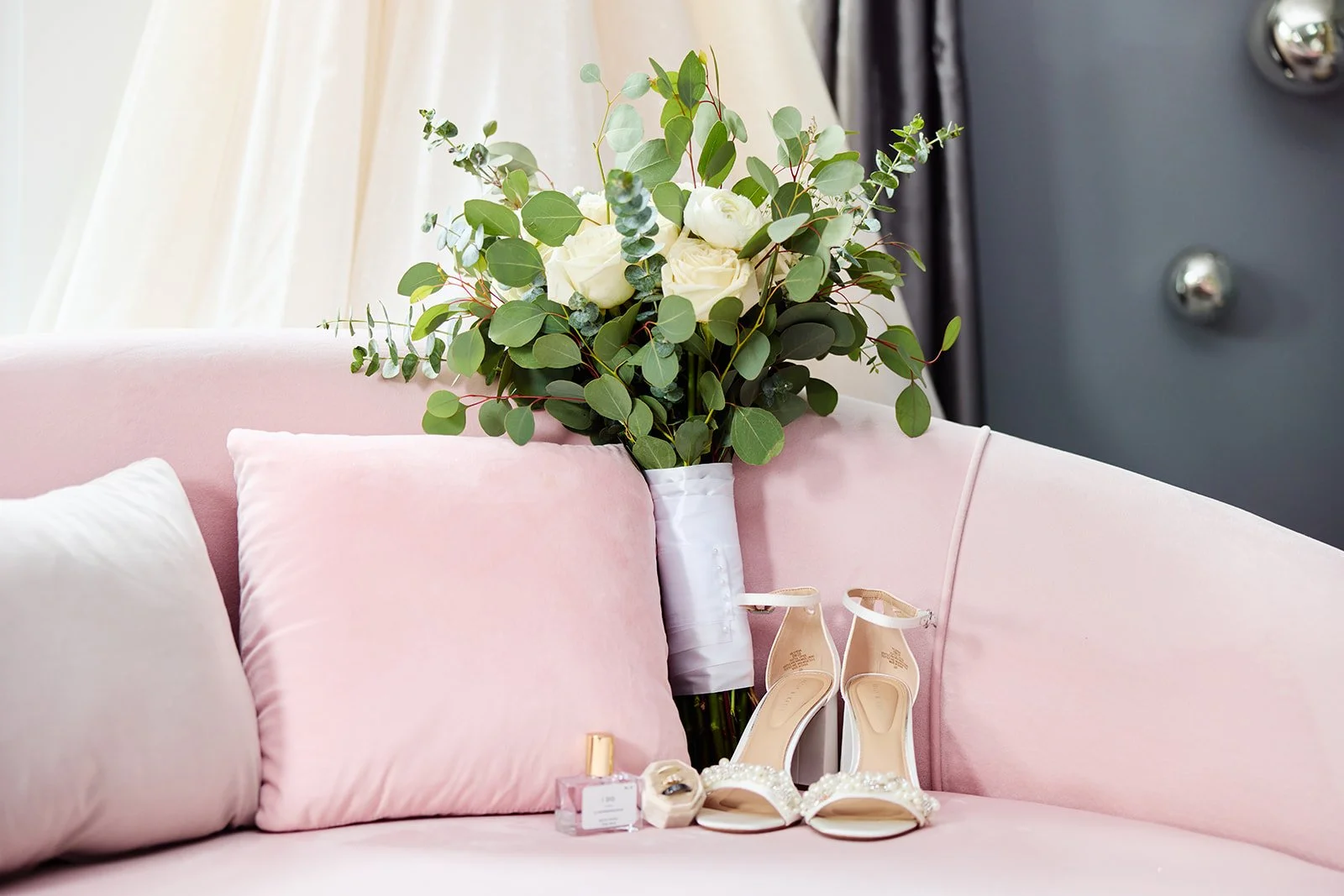 A bouquet of white roses and green foliage in a white vase on a pink velvet sofa with matching pillows, alongside white high-heeled sandals with pearls and a bottle of perfume.