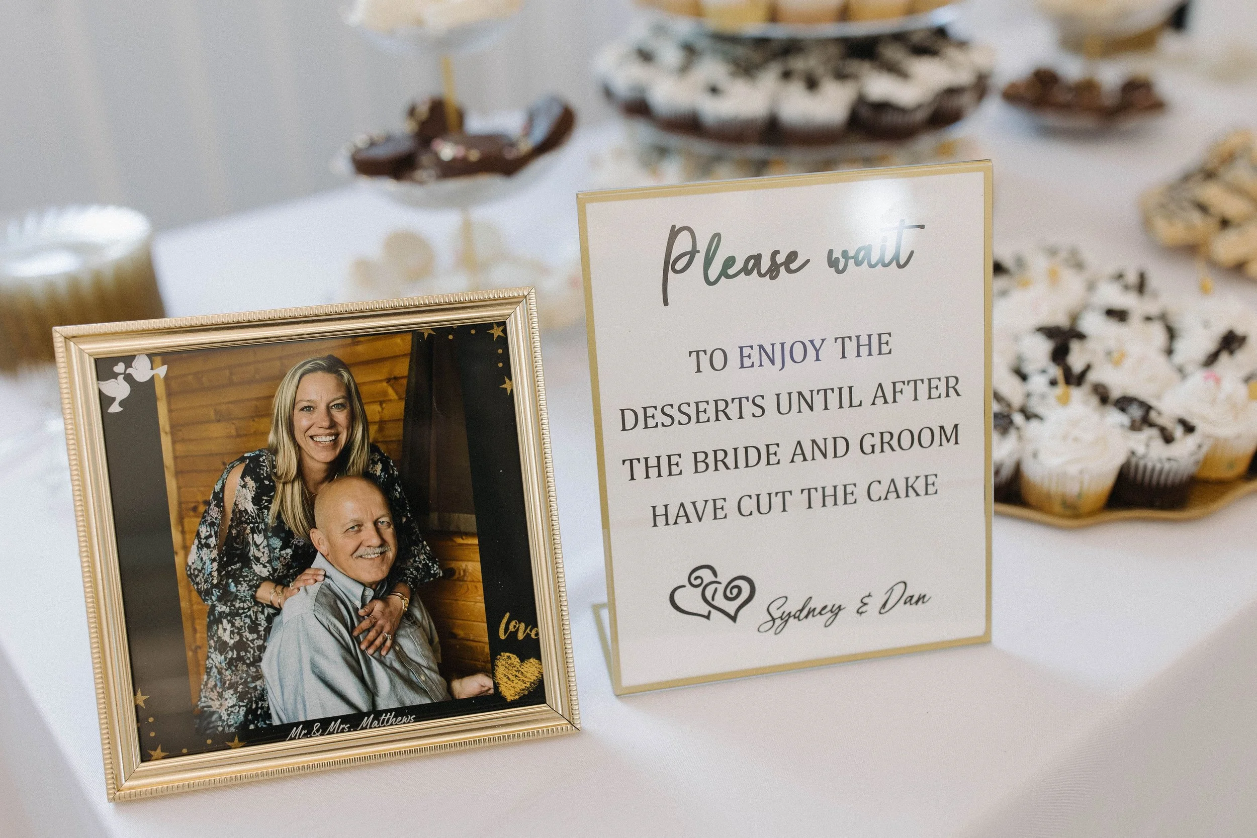 Wedding reception table with a framed photo of a smiling couple, a sign asking guests to wait to enjoy desserts until after the bride and groom cut the cake, and various desserts like cupcakes and cookies in the background.