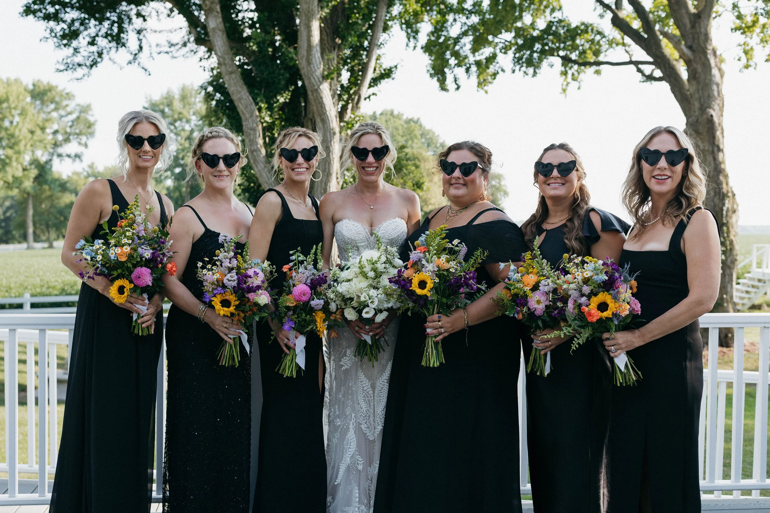 Group of women in black dresses with heart-shaped sunglasses holding colorful bouquets, standing outdoors on a sunny day with green trees in the background.