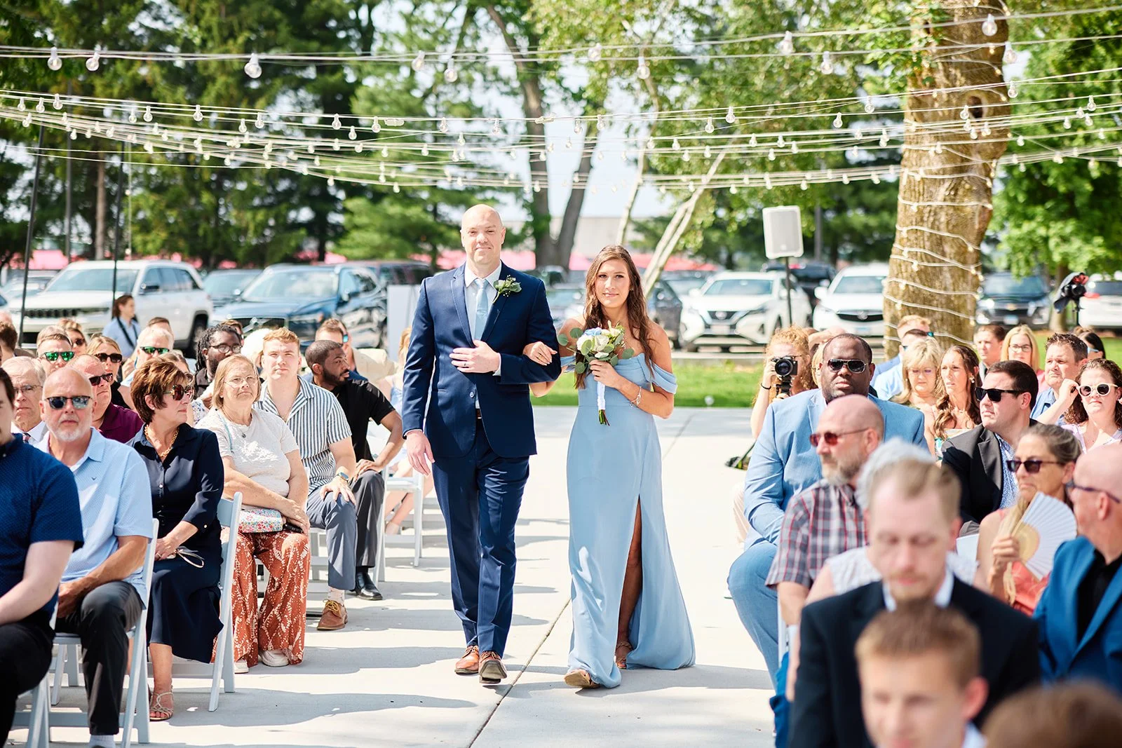 A wedding ceremony outdoors with the bride and groom walking down the aisle, surrounded by seated guests under string lights on a sunny day.