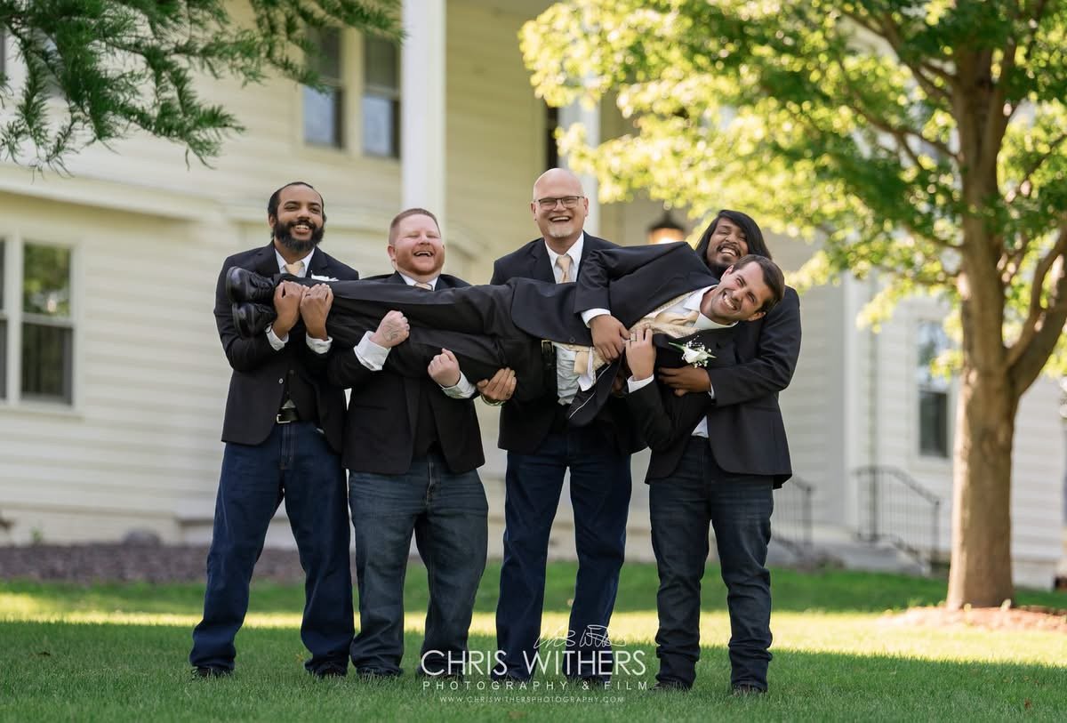 Five men in suits lifting a man in a tuxedo in the air outdoors near a house and trees.