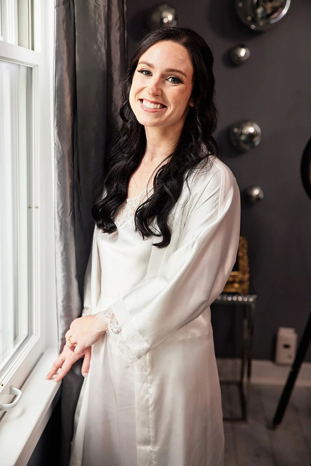 A woman with long black hair and light skin, smiling, standing by a window in a silky white robe with lace details at the cuffs, in a room with dark walls decorated with round metallic art pieces. A bride before she gets in her wedding dress. 