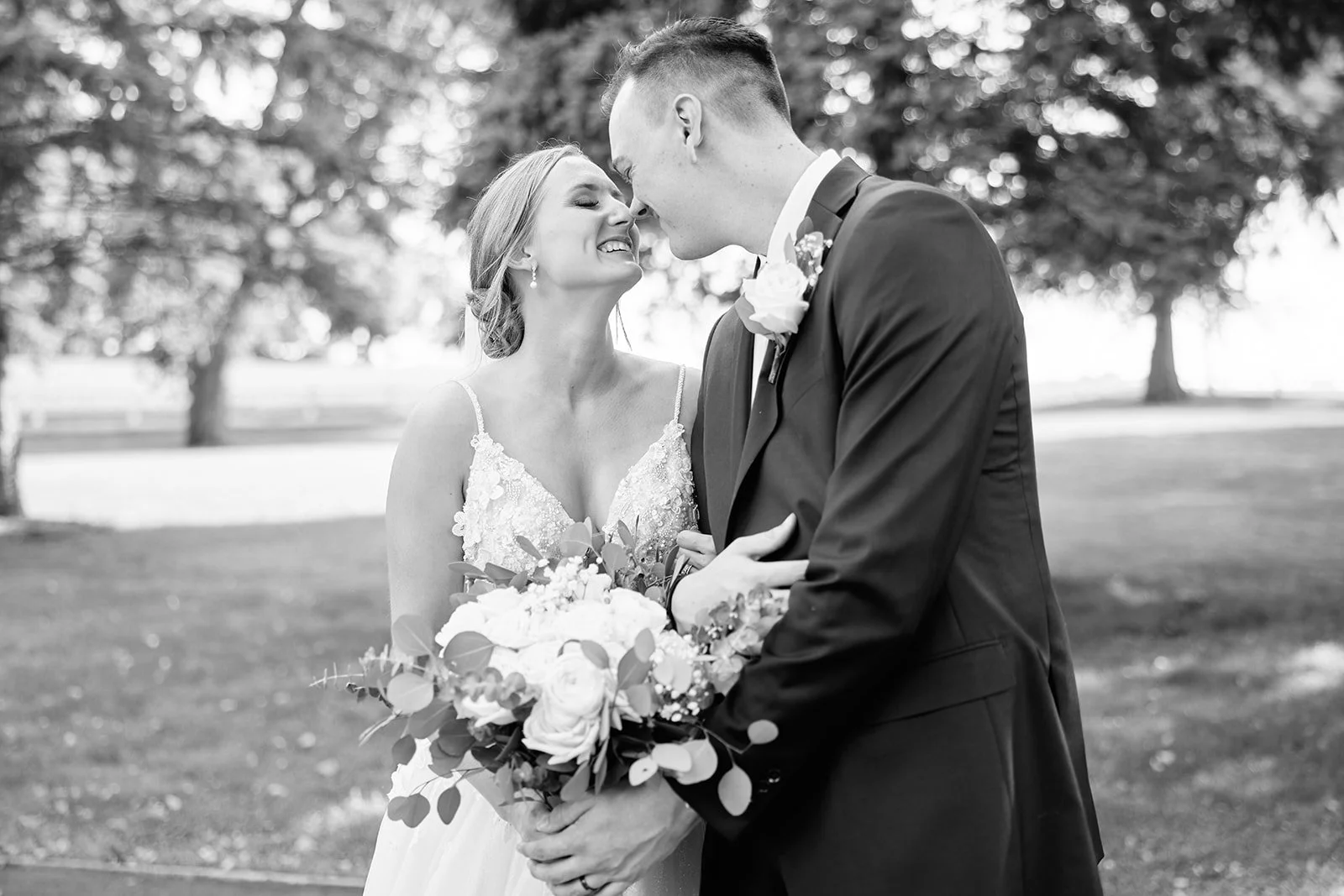Black and white photo of a bride and groom on their wedding day, standing close and smiling with noses touching outdoors, holding a bouquet of flowers.