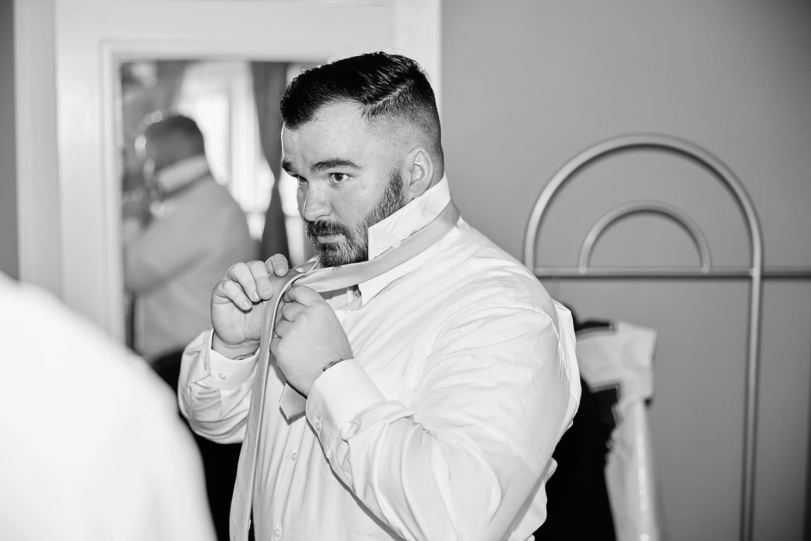 A man in a white dress shirt is tying a necktie in front of a mirror, with another man in the background also adjusting his attire.