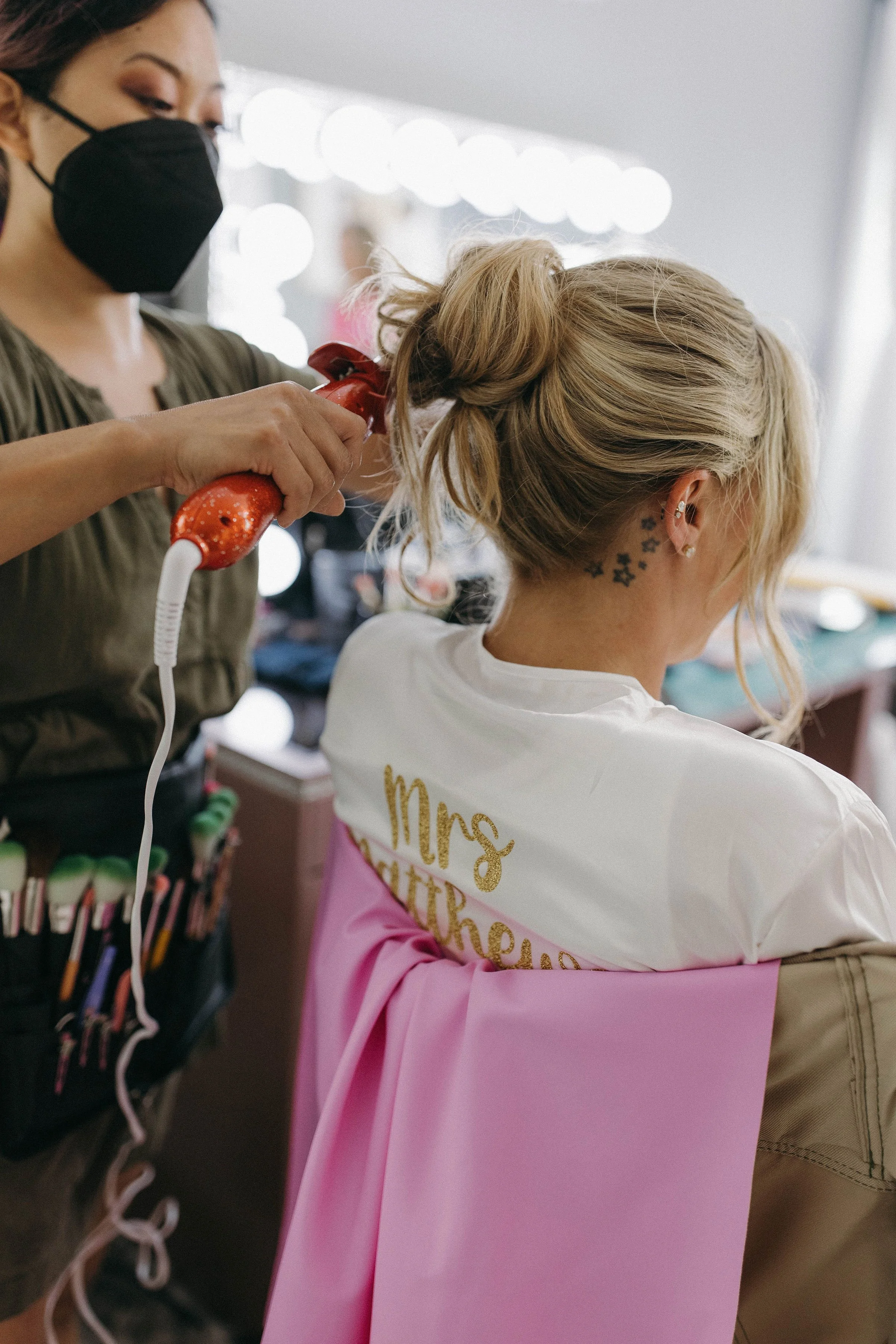 A woman is getting her blonde hair styled with a curling iron in a hair salon. She is wearing a white shirt with "Mrs" written on the back and a pink cape around her shoulders. The stylist, wearing a black face mask and apron with hair tools, is curl
