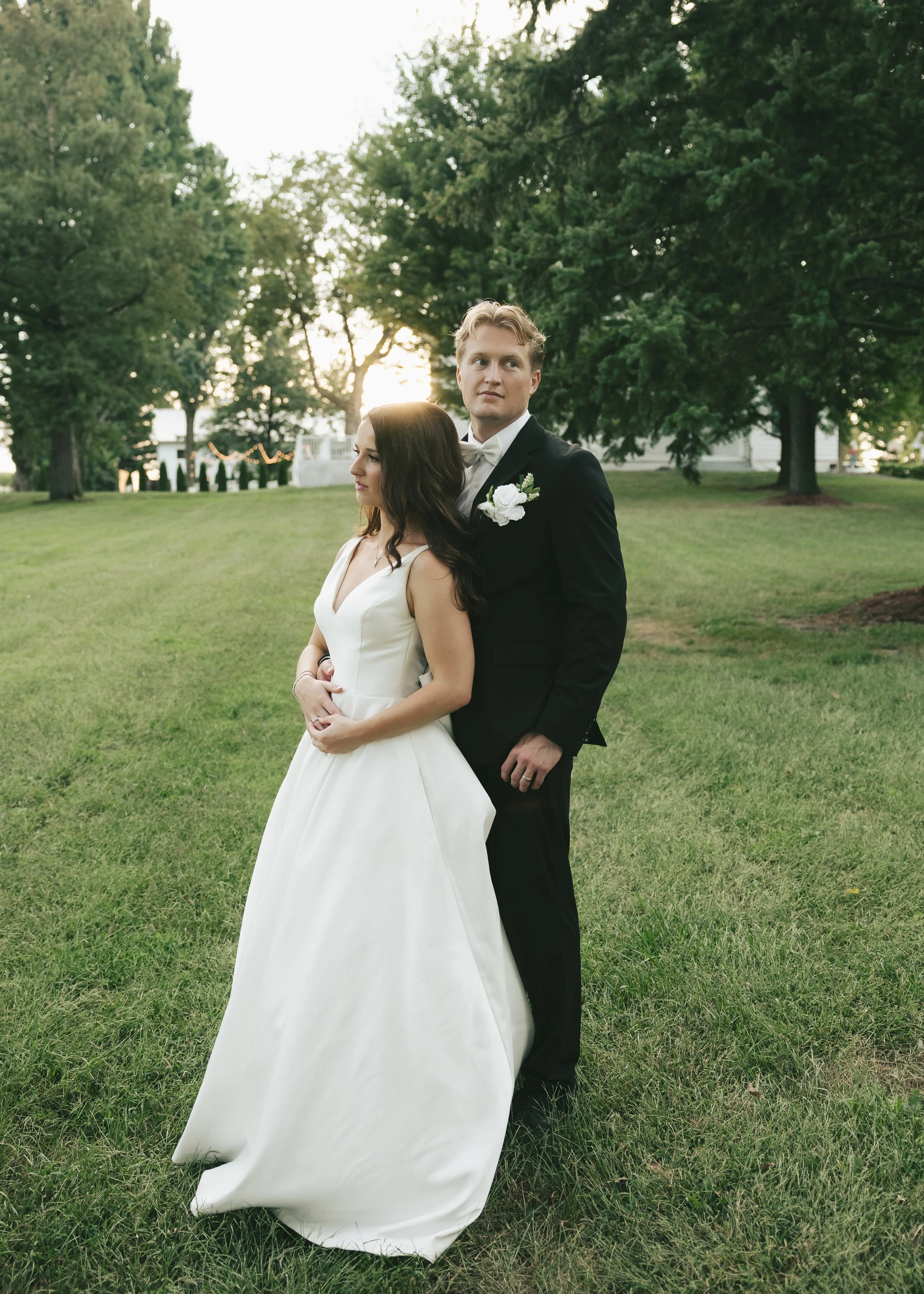A bride and groom standing outdoors on a grassy area with trees and a house in the background during sunset. The bride is wearing a white wedding gown and the groom is in a black tuxedo with a white bow tie and boutonniere.