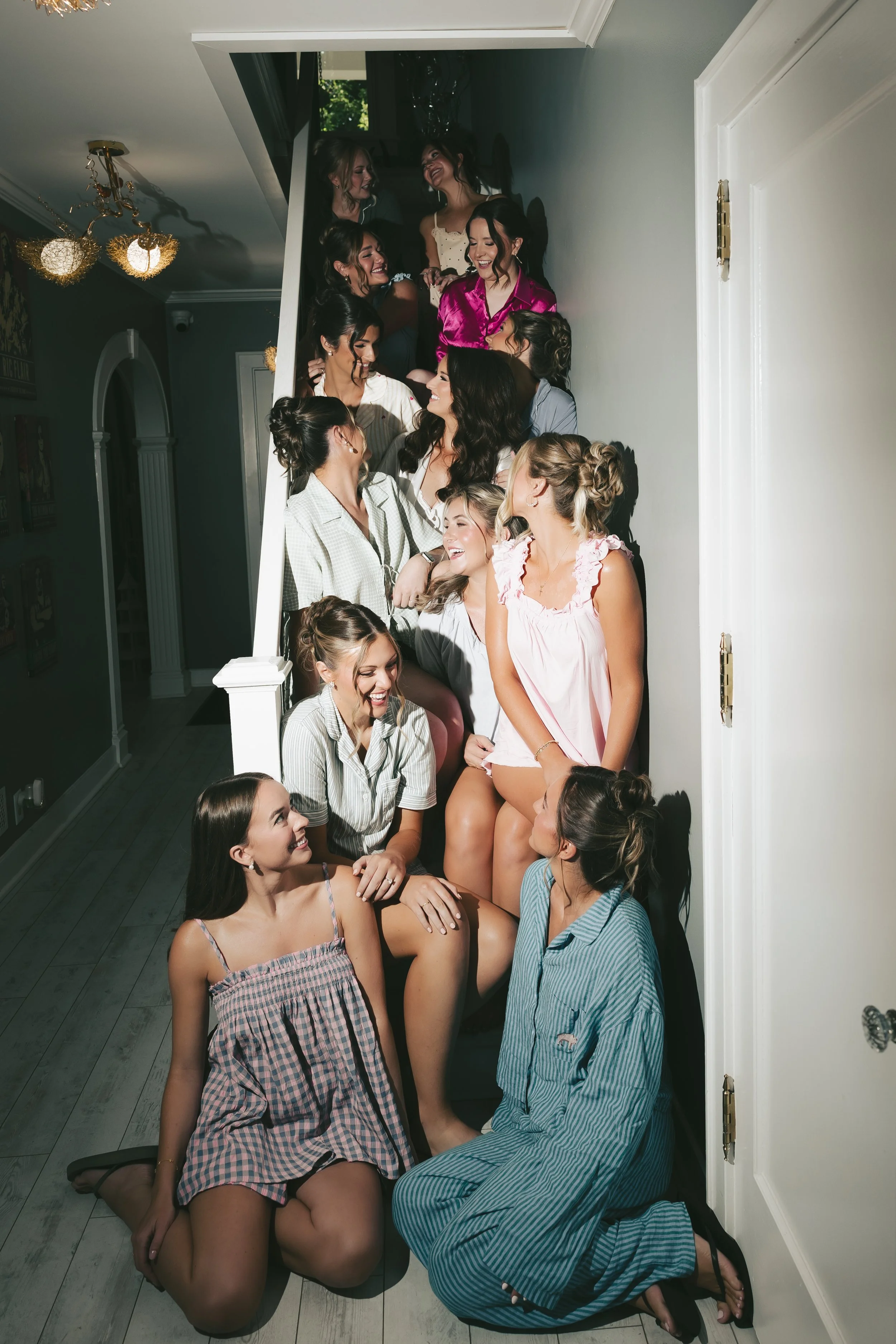 Group of women and a girl on a staircase, engaging in a candid moment, smiling and conversing, dressed in casual and sleepwear, in a well-lit room with decor and a chandelier.
