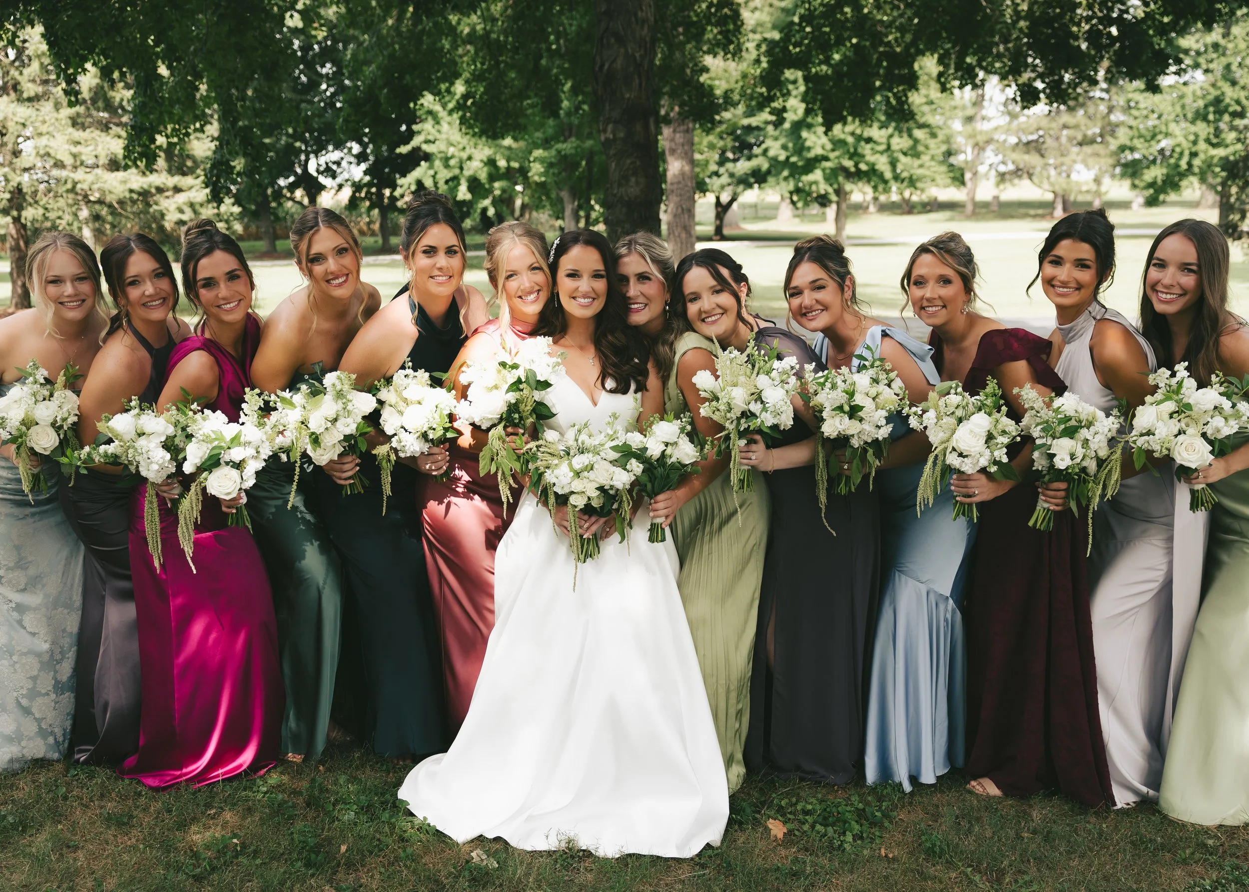 Group of women in colorful dresses gathered outdoors, holding white flower bouquets, smiling for a photo Pole Barn Chic with trees in the background, at a wedding venue in Central Illinois. 