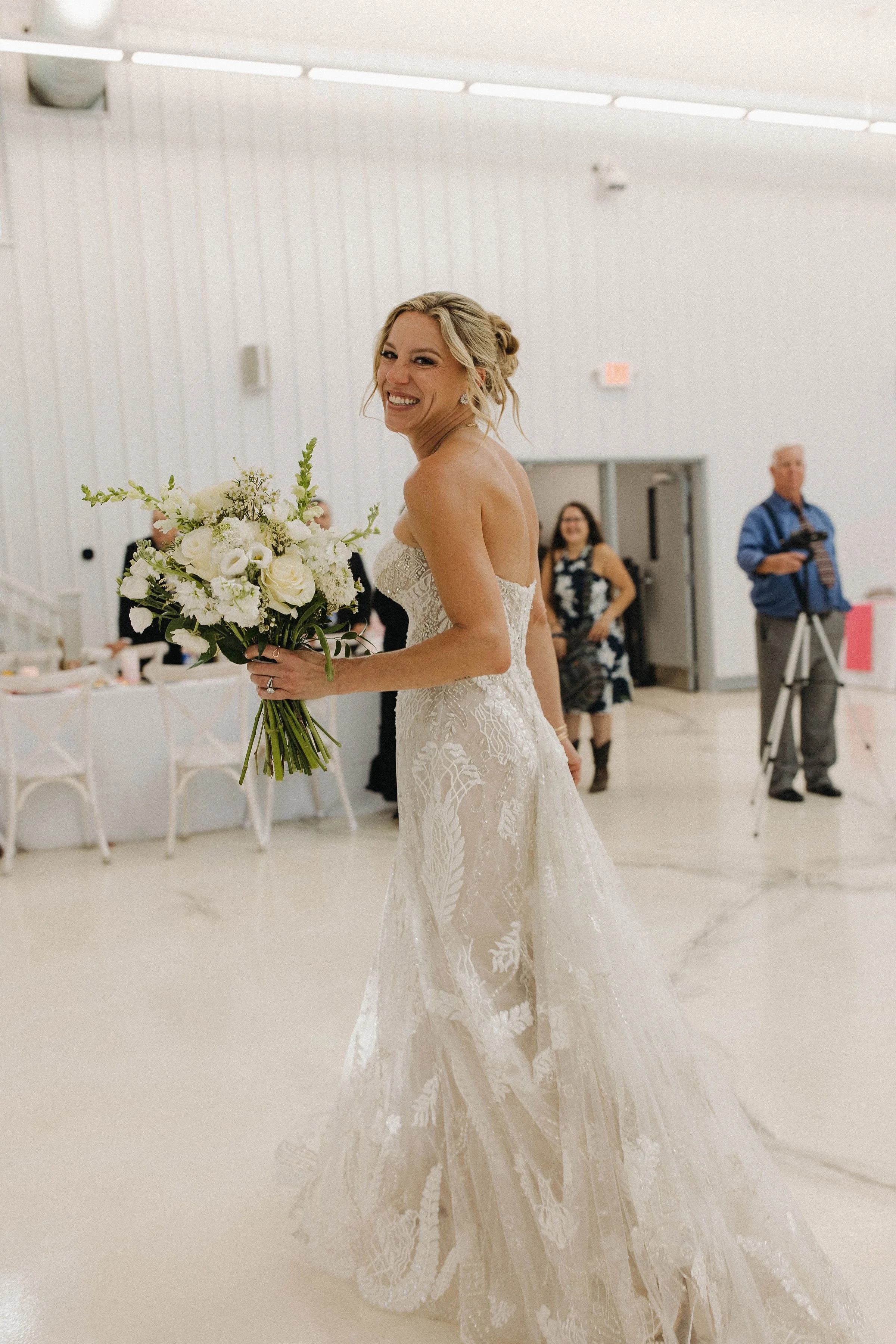 A joyful bride in a white lace wedding gown holds a bouquet of white flowers, smiling at a reception hall.