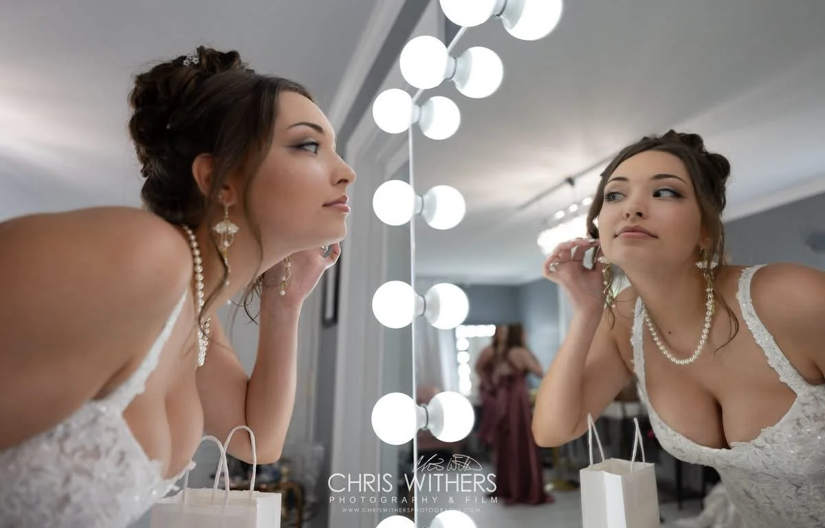 Bridal bride getting ready, looking in the mirror, wearing a white lace wedding dress, pearl necklace, earrings, and a tiara with her hair styled up, in a well-lit room with a mirror decorated with round lights.