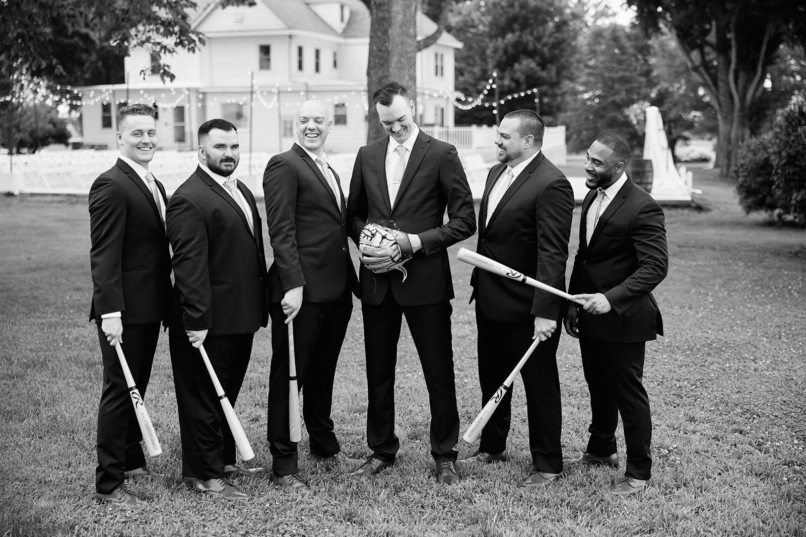 Seven men in suits standing on grass, holding baseball bats and a baseball glove, smiling, with a house and string lights in the background.