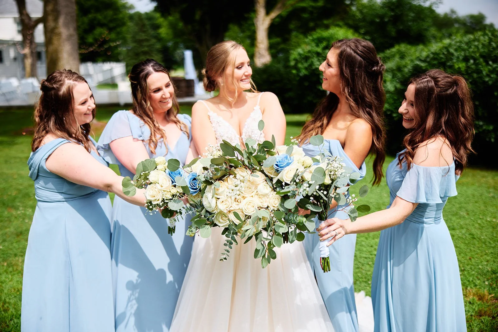 A bride in a white wedding dress holding a large bouquet of white and blue roses, surrounded by five bridesmaids in matching light blue dresses, smiling and looking at each other outdoors on a grassy area with trees in the background.