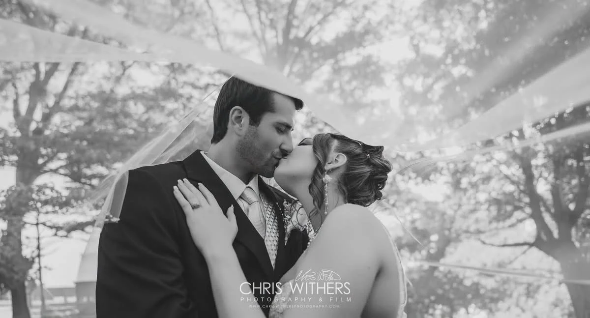 A black and white photograph of a bride and groom kissing under a veil outdoors, with trees in the background.