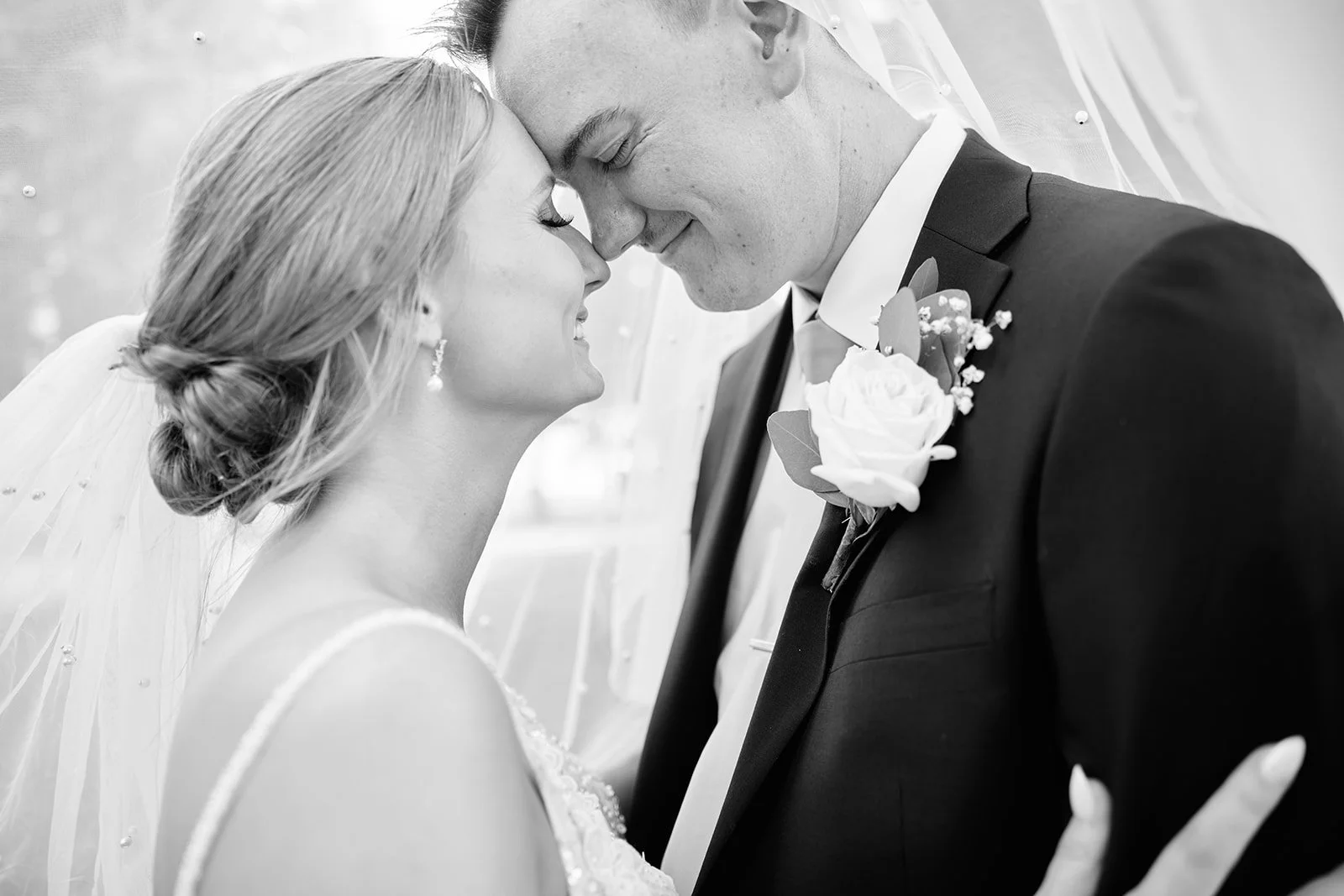 Black and white photo of a bride and groom with foreheads touching, smiling, and close eyes, during their wedding.