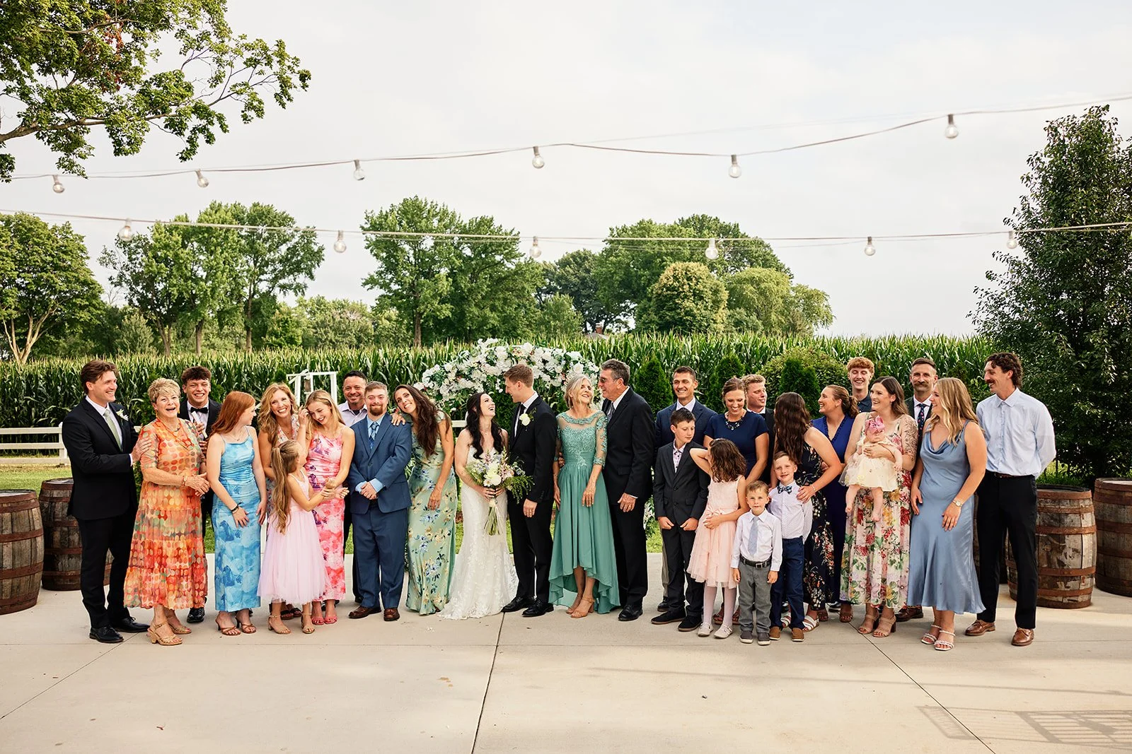 A large group of people gathered outdoors for a wedding celebration, with a bride and groom standing in the center, surrounded by family and friends, all dressed in formal and semi-formal attire, with a lush green background and string lights overhea