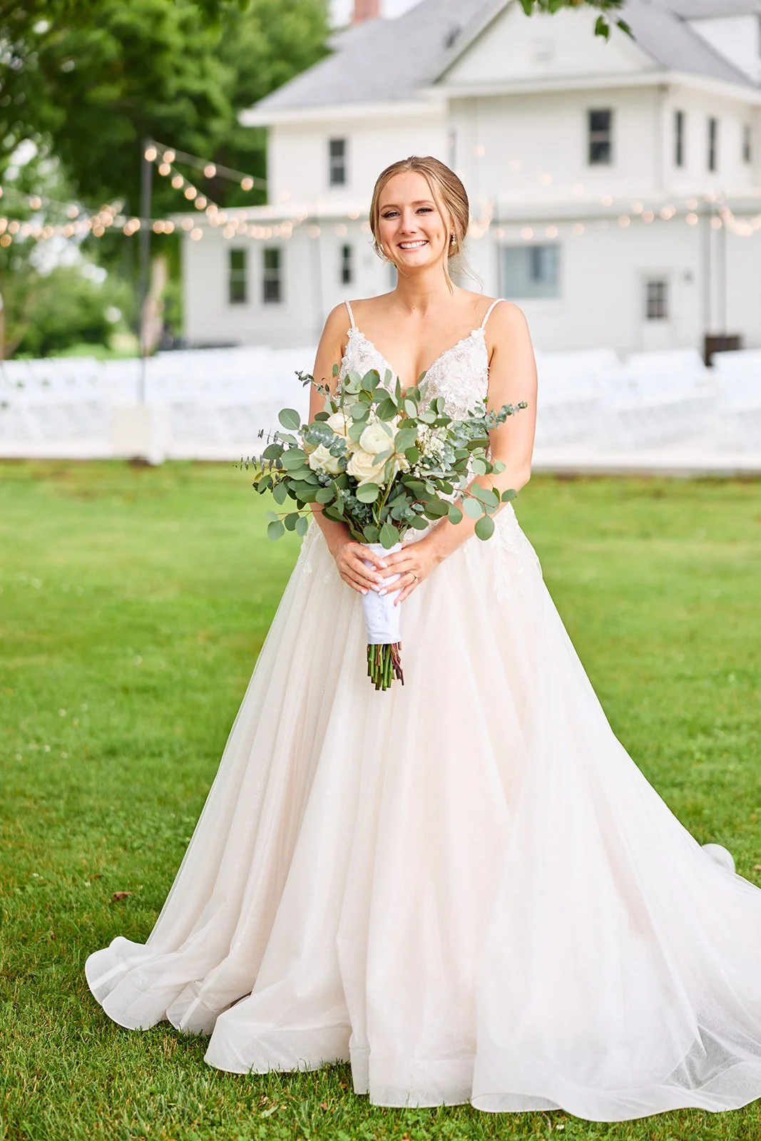A bride in a white wedding gown holding a bouquet of white roses and greenery, standing on a green lawn with a house and string lights in the background.