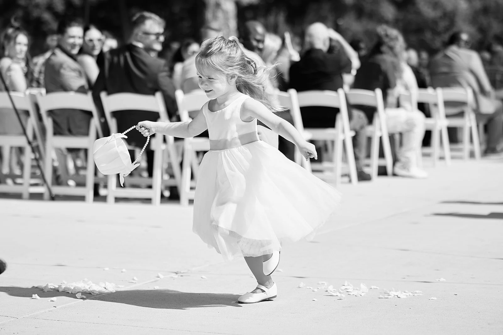 A young flower girl in a white dress twirling at an outdoor wedding ceremony. Guests seated in the background.