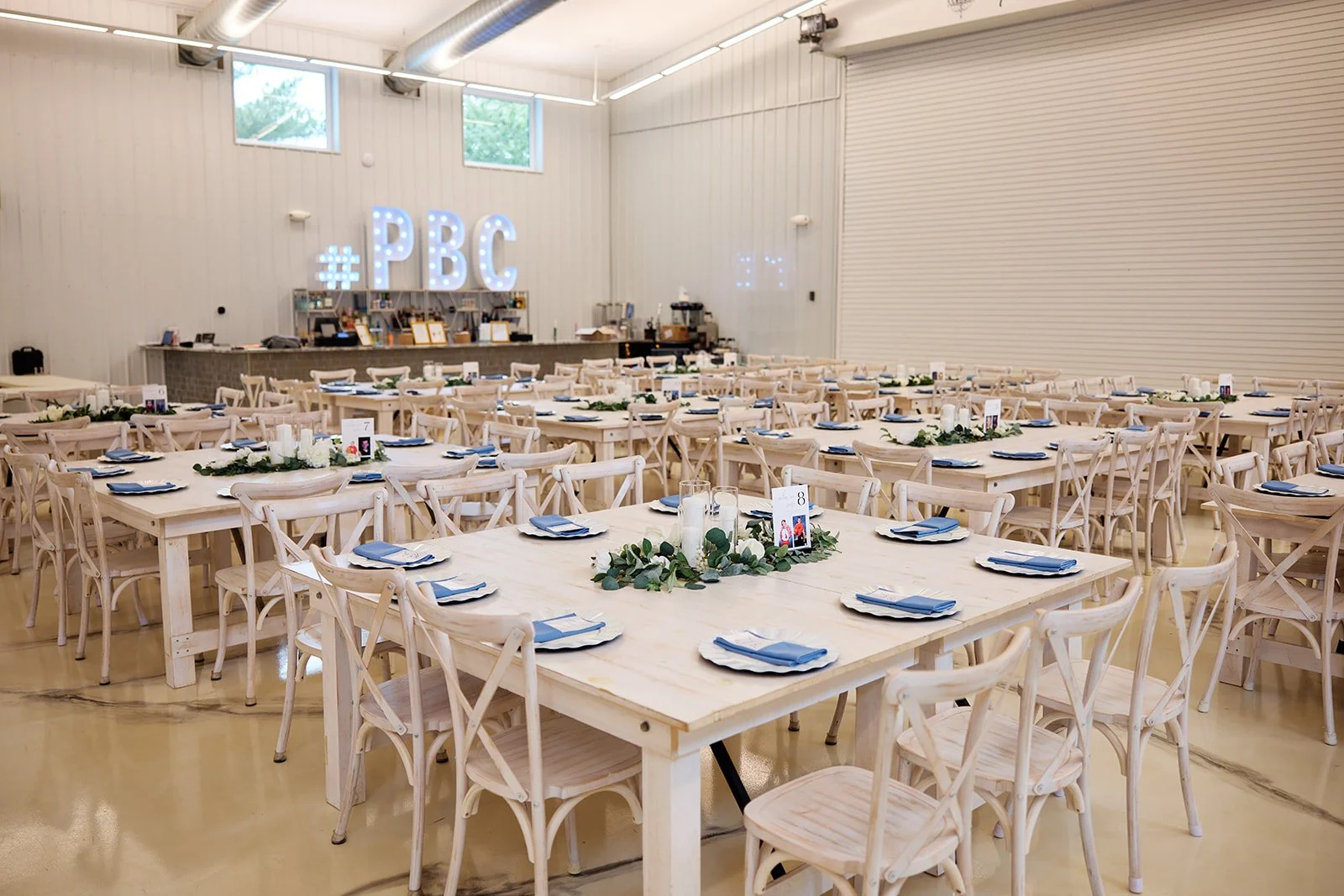 Large indoor space decorated for an event with many light wood tables and chairs. The tables are set with white plates, blue napkins, and centerpieces of candles and greenery. Elevated signage in the background displays '#PBC' with glowing letters.