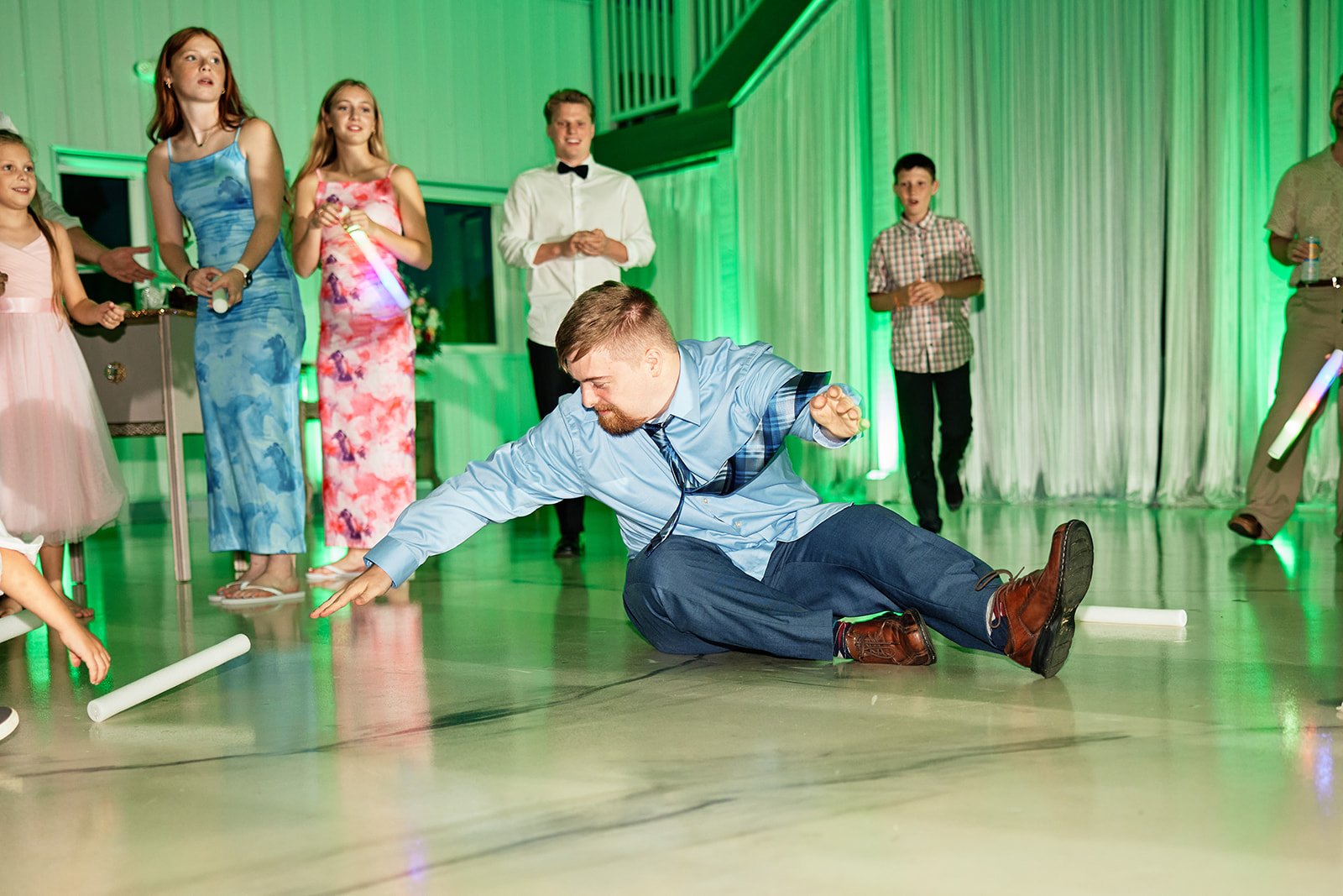 A man break dancing on the dance floor during a party game, surrounded by children and teenagers, with some holding glow sticks and others watching, in a decorated room with green lighting.