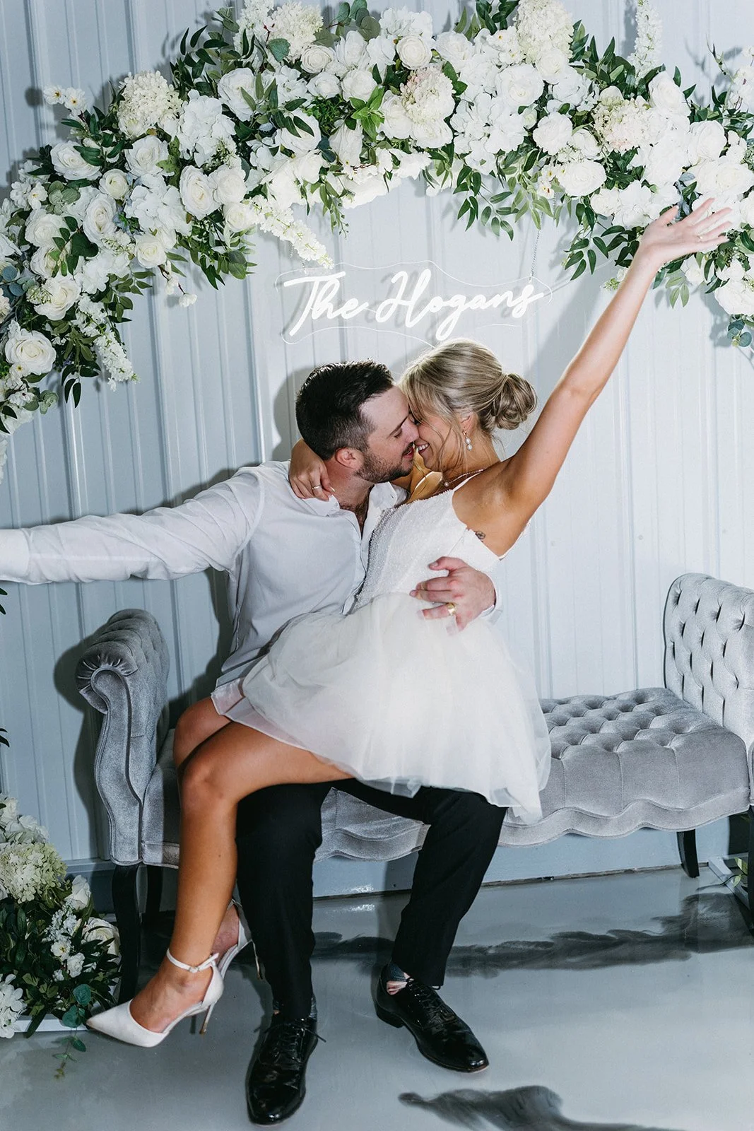 A couple celebrating a wedding, sitting on a vintage gray velvet sofa, surrounded by white flowers and greenery, with a neon sign saying "The Hogans" behind them. The woman is in a white dress, with her arms raised, and the man is in a white shirt an