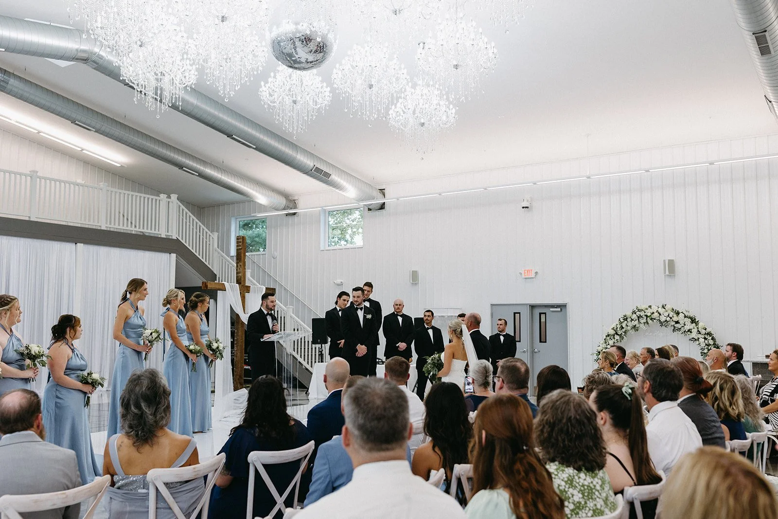 Wedding ceremony in the PBC Venue a white indoor venue with guests seated, bridal party standing, a bride and groom exchanging vows, and a large floral arch in the background.