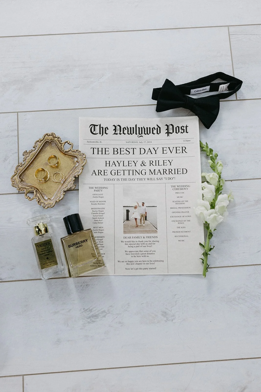 Wedding invitation and accessories including a black bow tie, a floral hairpiece, two perfume bottles, and wedding rings on a decorative tray.